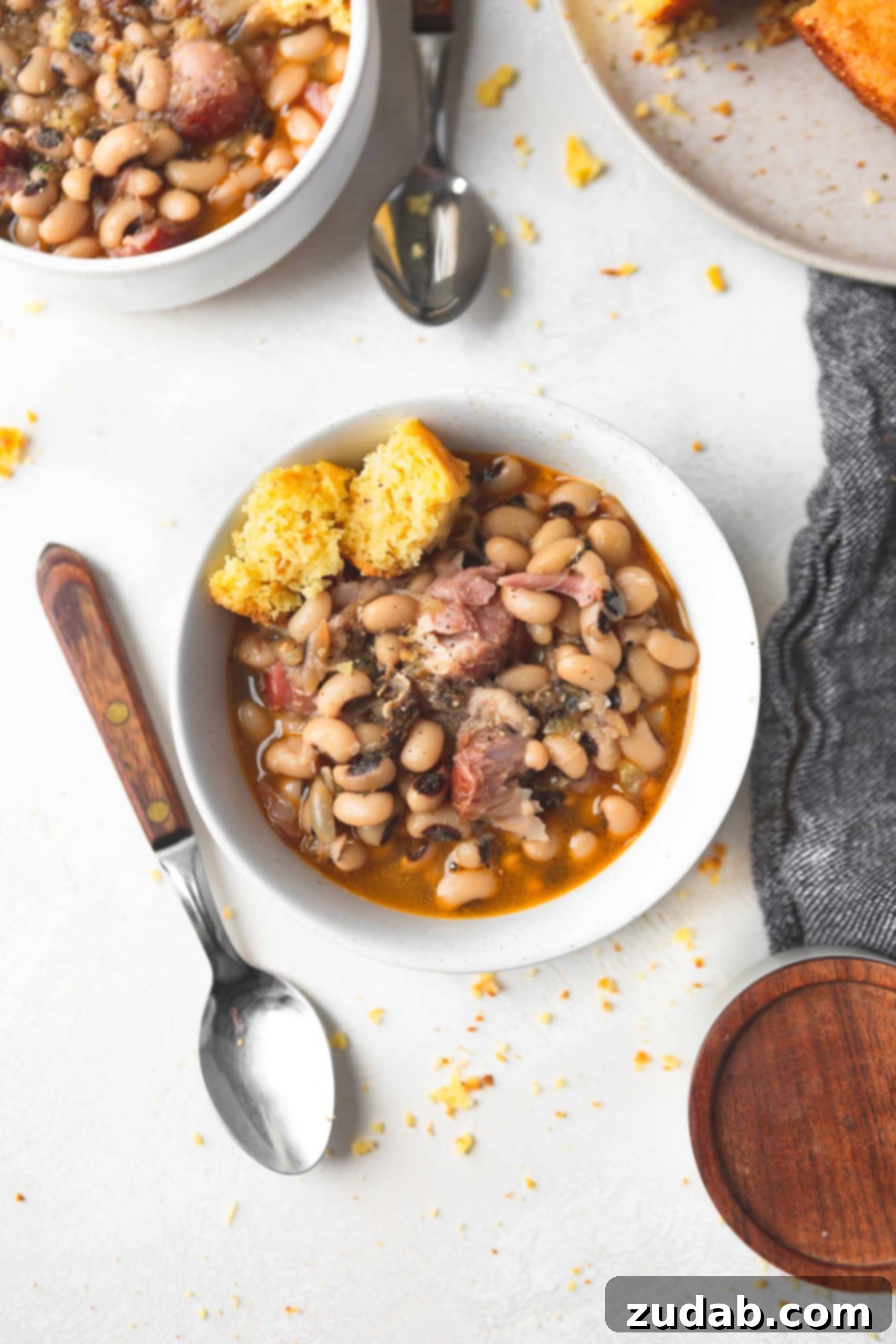 A bowl of southern black eyed peas with chunks of ham and a broken piece of corn bread. A spoon is next to the bowl of black eyed peas.