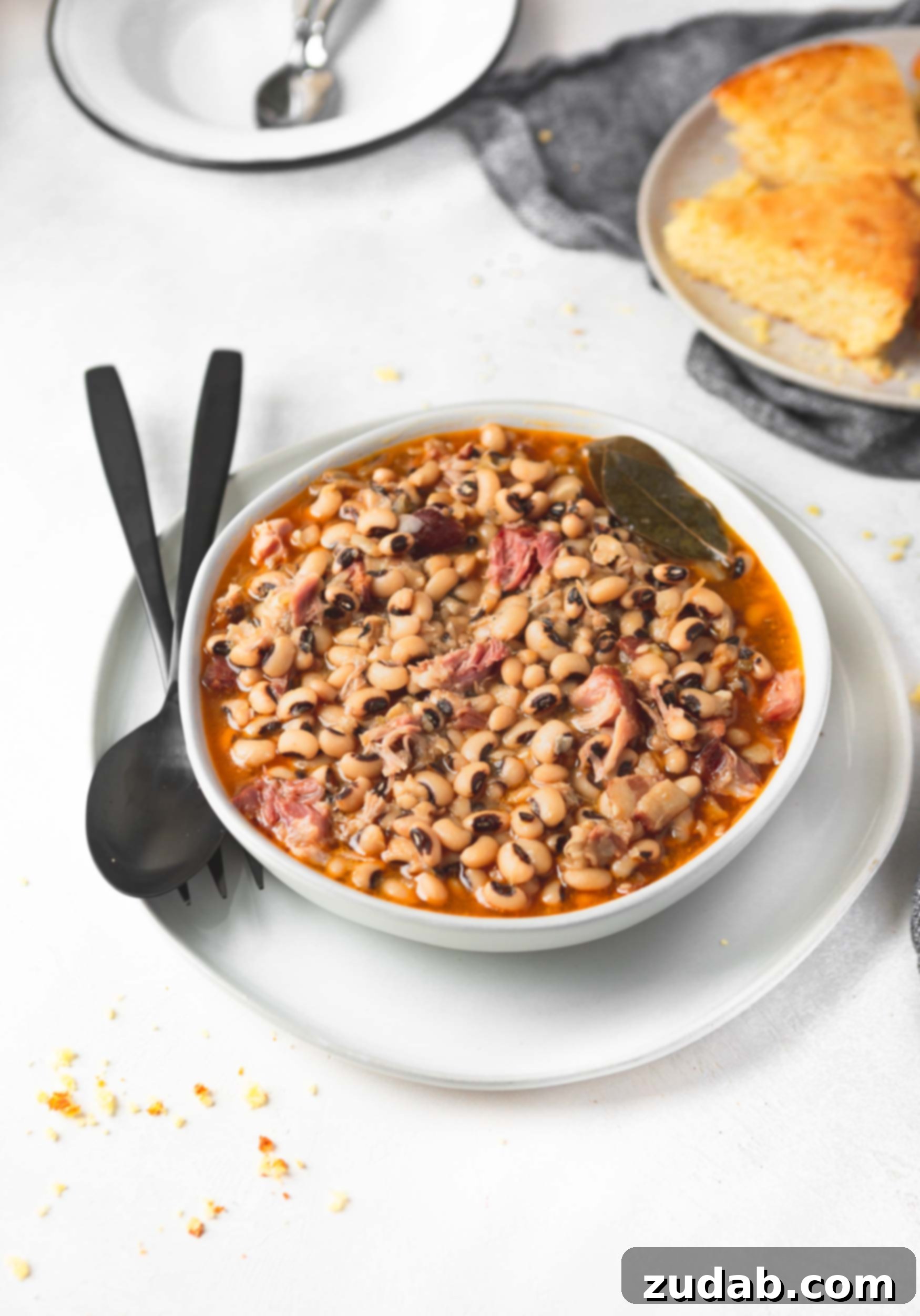 A bowl of black eyed peas. The bowl is sitting on a saucer with a fork and spoon next to it. There is a plate of cornbread in the background.