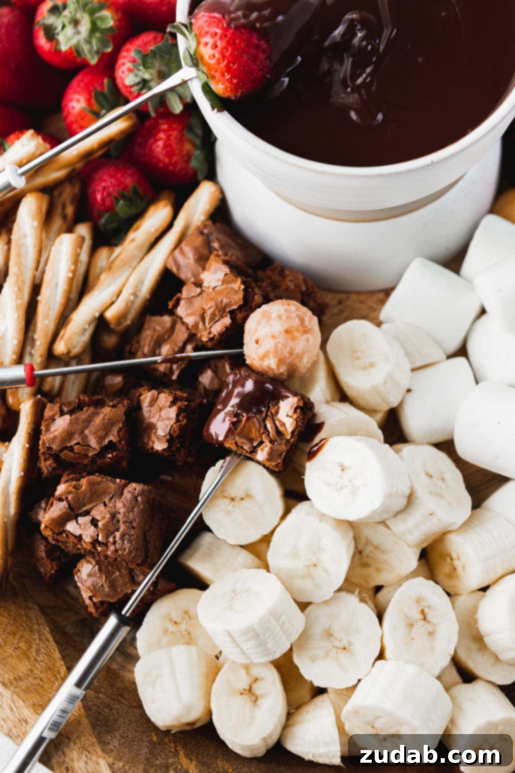 Chocolate Fondue A vibrant and inviting spread of various dippers surrounding a bowl of rich chocolate fondue. The image features fresh, juicy strawberries, sliced bananas, indulgent brownie bites, crispy pretzels, and fluffy marshmallows, all perfectly arranged and ready for dipping.