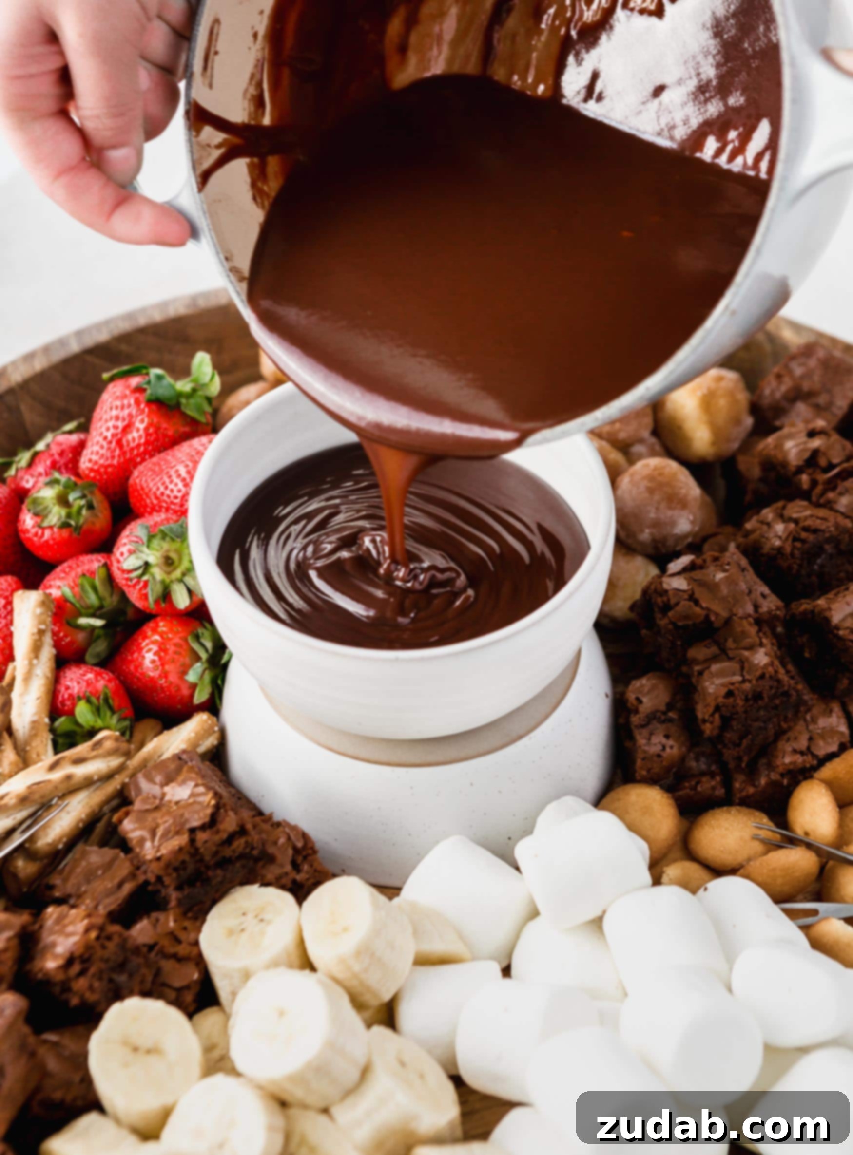 Chocolate Fondue Rich, glossy chocolate fondue being gracefully poured from a saucepan into an elegant serving bowl. The bowl is positioned on a rustic tray, which is artfully surrounded by an array of delightful dippers, including juicy strawberries, fluffy marshmallows, assorted cookies, and crunchy nuts, all poised for a decadent dipping experience.