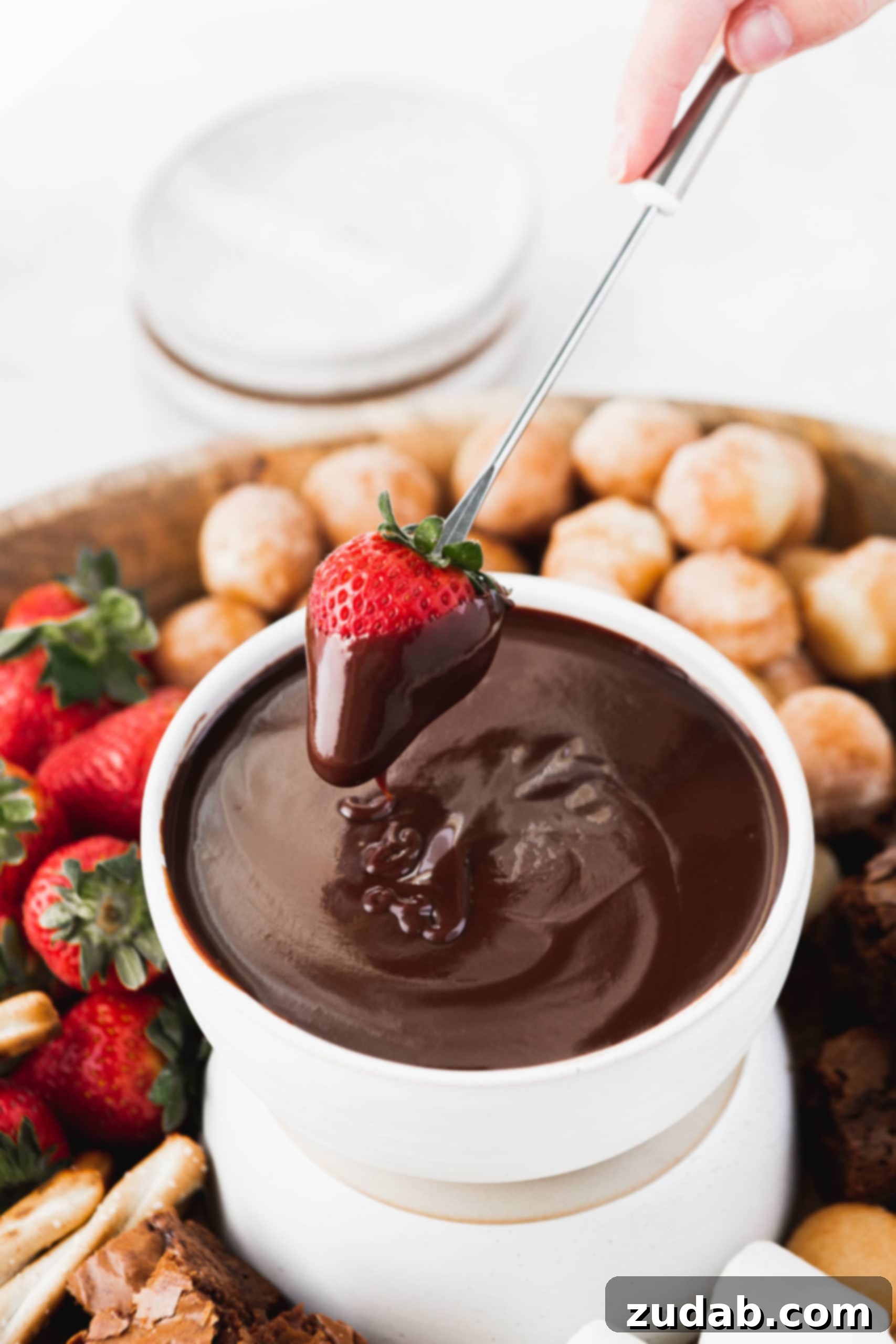 Chocolate Fondue A bowl of luscious, dark chocolate fondue takes center stage, with a glistening strawberry that has just been dipped, its surface coated in rich chocolate. In the softly blurred background, an enticing spread of more fresh strawberries, indulgent brownies, and sweet donut holes awaits, promising endless dipping pleasure.