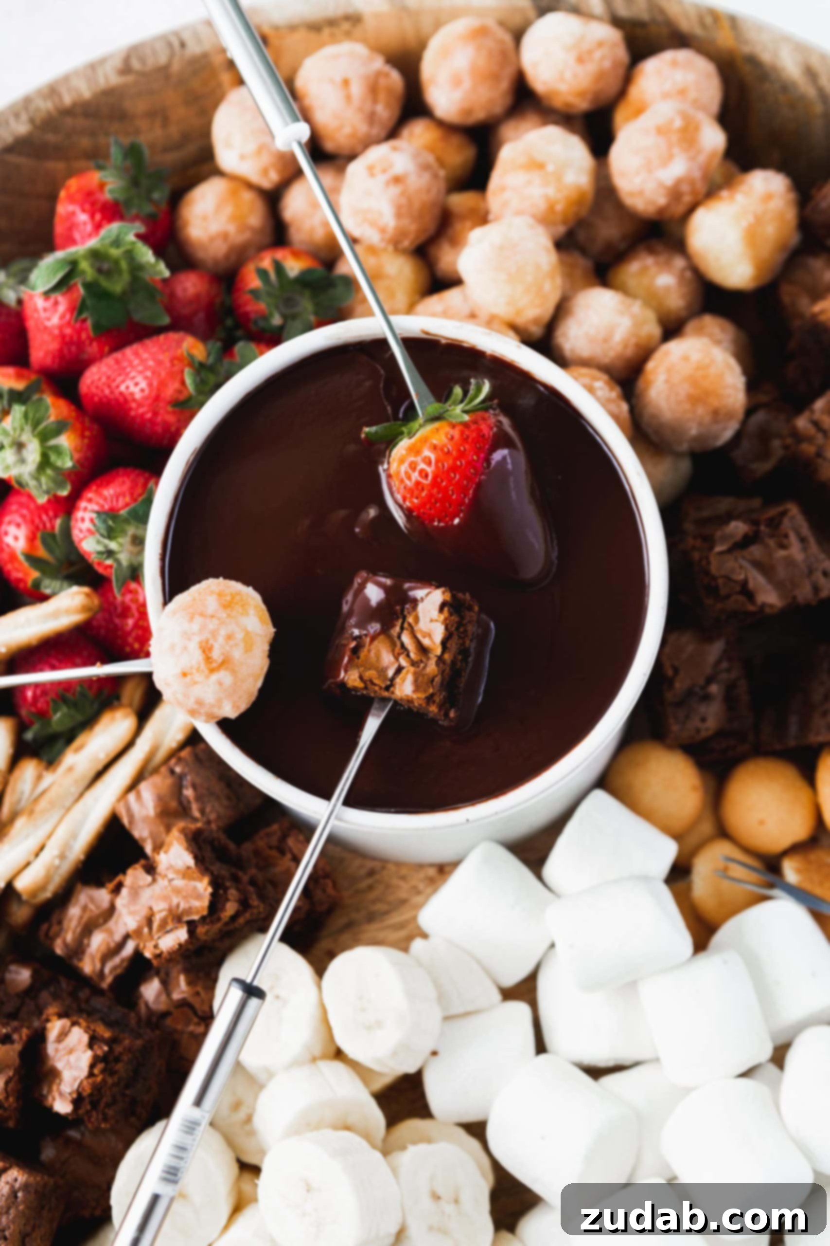 Chocolate Fondue A lively and inviting scene featuring a central bowl of glistening chocolate fondue. Multiple hands are busy dipping a strawberry, a brownie, and a donut hole into the rich, melted chocolate, highlighting the interactive and communal joy of the dessert. The fondue pot is surrounded by an abundant and colorful assortment of dippable treats, including more fresh strawberries, sweet donut holes, fudgy brownies, soft marshmallows, and a variety of other delicious items.