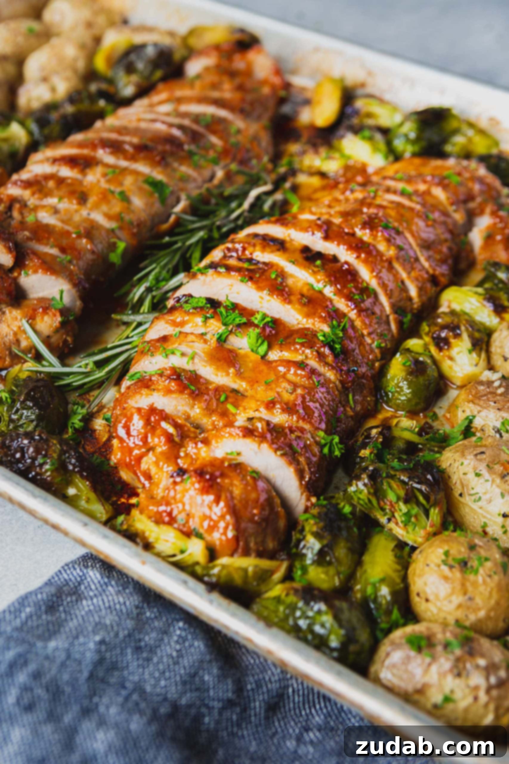 A Picture of two roasted pork loins that are browned on top and sliced for serving. You can see herbs and roasted potatoes on the pan next to the pork loin.