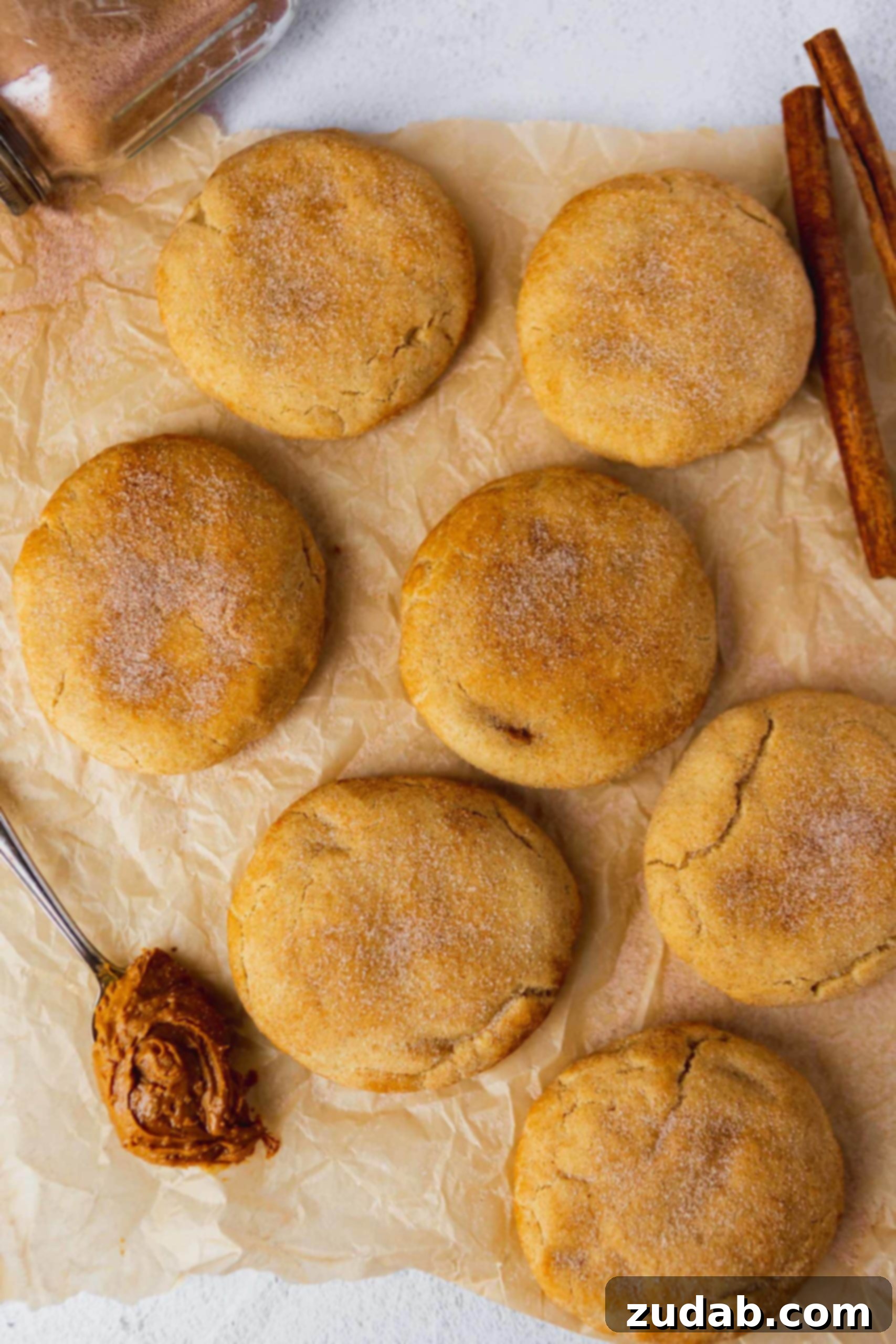 Biscoff Stuffed Snickerdoodle Cookies Baked large and thick snickerdoodles on parchment paper. They are sprinkled with cinnamon sugar and a spoonful of biscoff is next to the cookies.