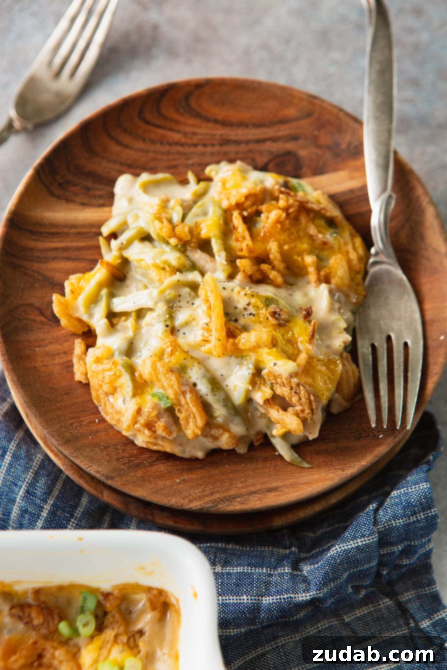 Ultimate Classic Green Bean Casserole 2 A serving of creamy green bean casserole on a rustic wooden plate with a fork beside it. The casserole is generously topped with golden brown, crunchy French fried onions, and you can see the vibrant green beans mixed within the rich sauce.