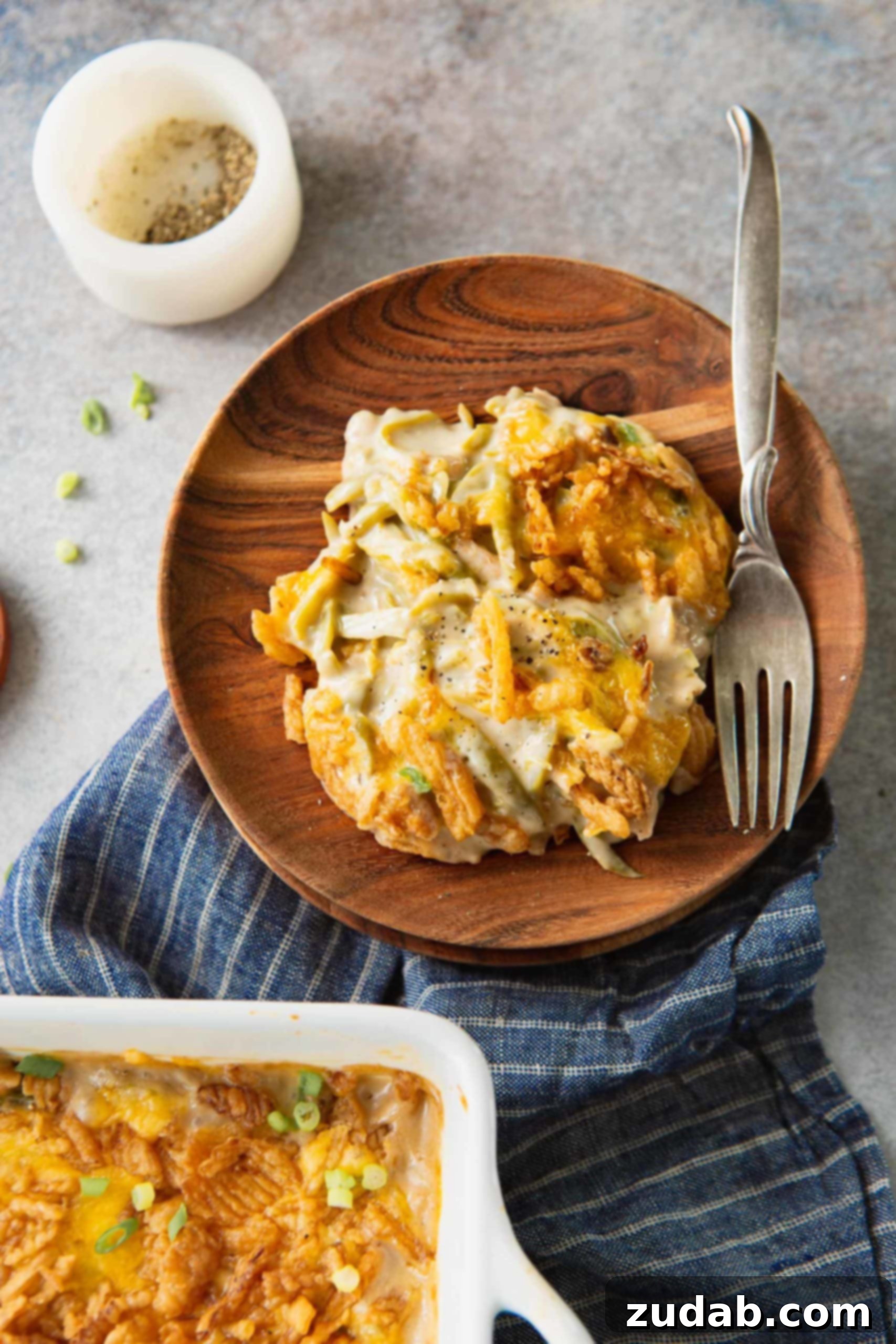 Ultimate Classic Green Bean Casserole 4 A serving of classic green bean casserole on a rustic wooden plate, with a fork resting beside it. The casserole is rich and creamy, featuring tender green beans and a generous, crispy topping of golden French fried onions, highlighting its comforting appeal.