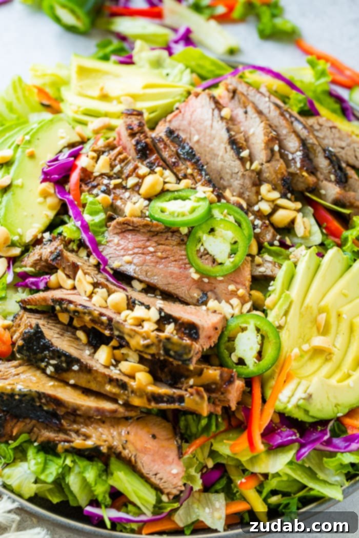 Over 25 Healthy Recipes 16 A grey plate piled with mixed greens, colorful vegetables, and marinated Thai peanut steak, drizzled with a vibrant ginger dressing and sesame seeds.