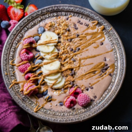 Over 25 Healthy Recipes 3 A vibrant chocolate banana smoothie bowl adorned with raspberries, banana slices, blueberries, granola, and chocolate chips, drizzled with peanut butter. A glass of milk sits in the background.