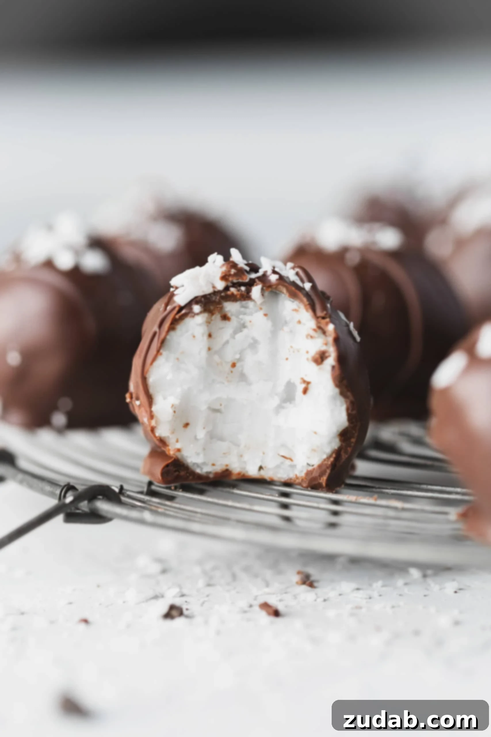 Chocolate Covered Coconut Balls A chocolate covered coconut ball that has been broken in half so you can see the creamy white coconut center. There are other coconut balls in the background.