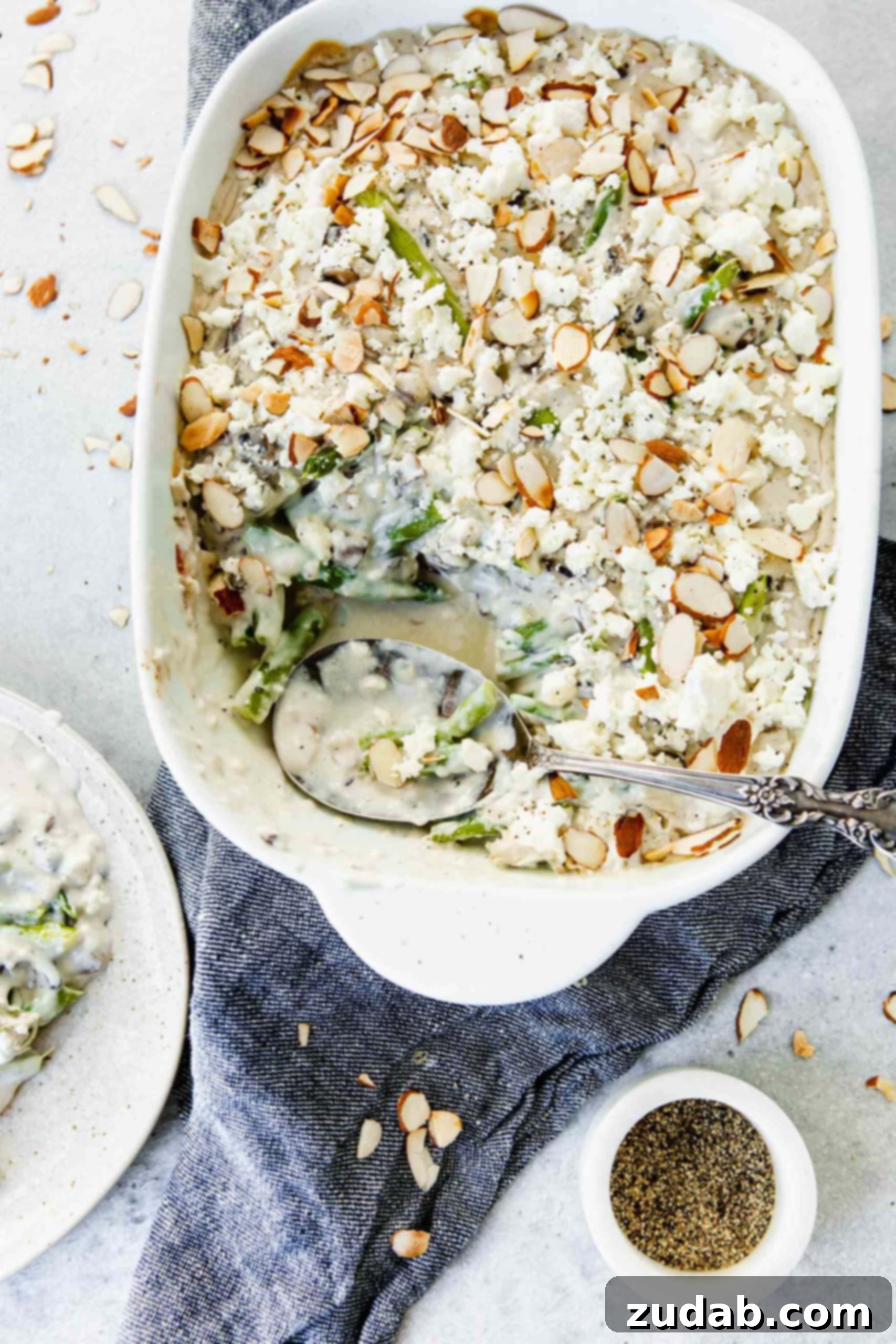 Feta Green Bean Casserole A casserole dish filled with vibrant green beans, creamy feta cheese sauce, and sliced almonds on top. A serving spoon is in the dish, showcasing the rich textures. A dinner plate with a spoonful of the casserole is nearby, ready to be enjoyed.