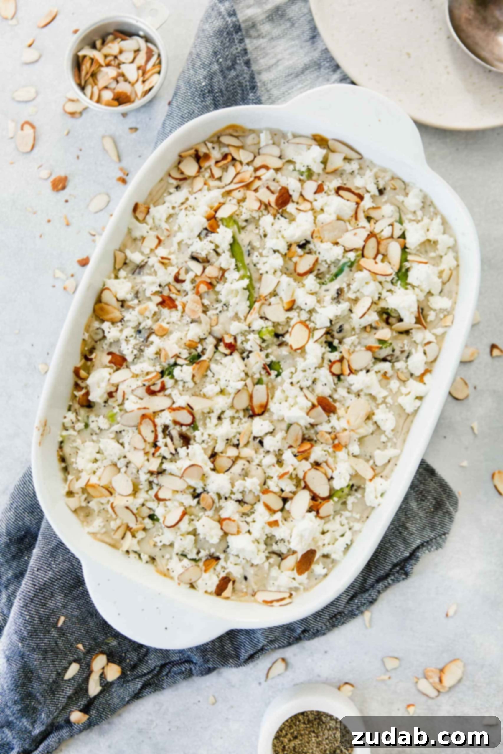 Feta Green Bean Casserole A close-up view of Feta Green Bean Casserole in a baking dish. The green beans are generously coated in a luscious white cream sauce, topped with crumbled feta cheese and perfectly golden sliced almonds.