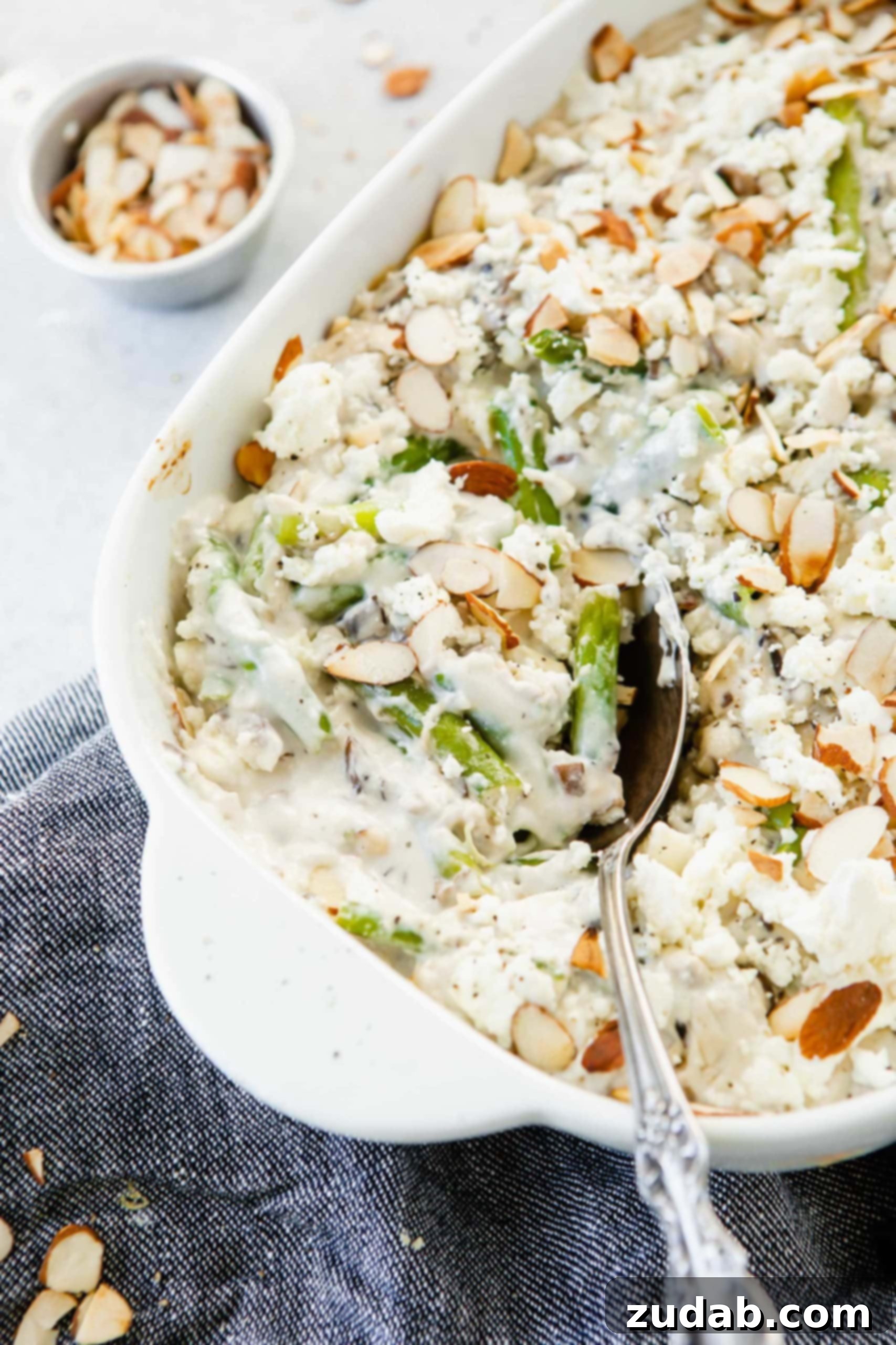 Feta Green Bean Casserole A close-up shot of the Feta Green Bean Casserole in a serving dish, with a serving spoon poised to scoop. The creamy white sauce beautifully coats the green beans, and a generous sprinkle of sliced almonds sits atop, promising both flavor and texture.