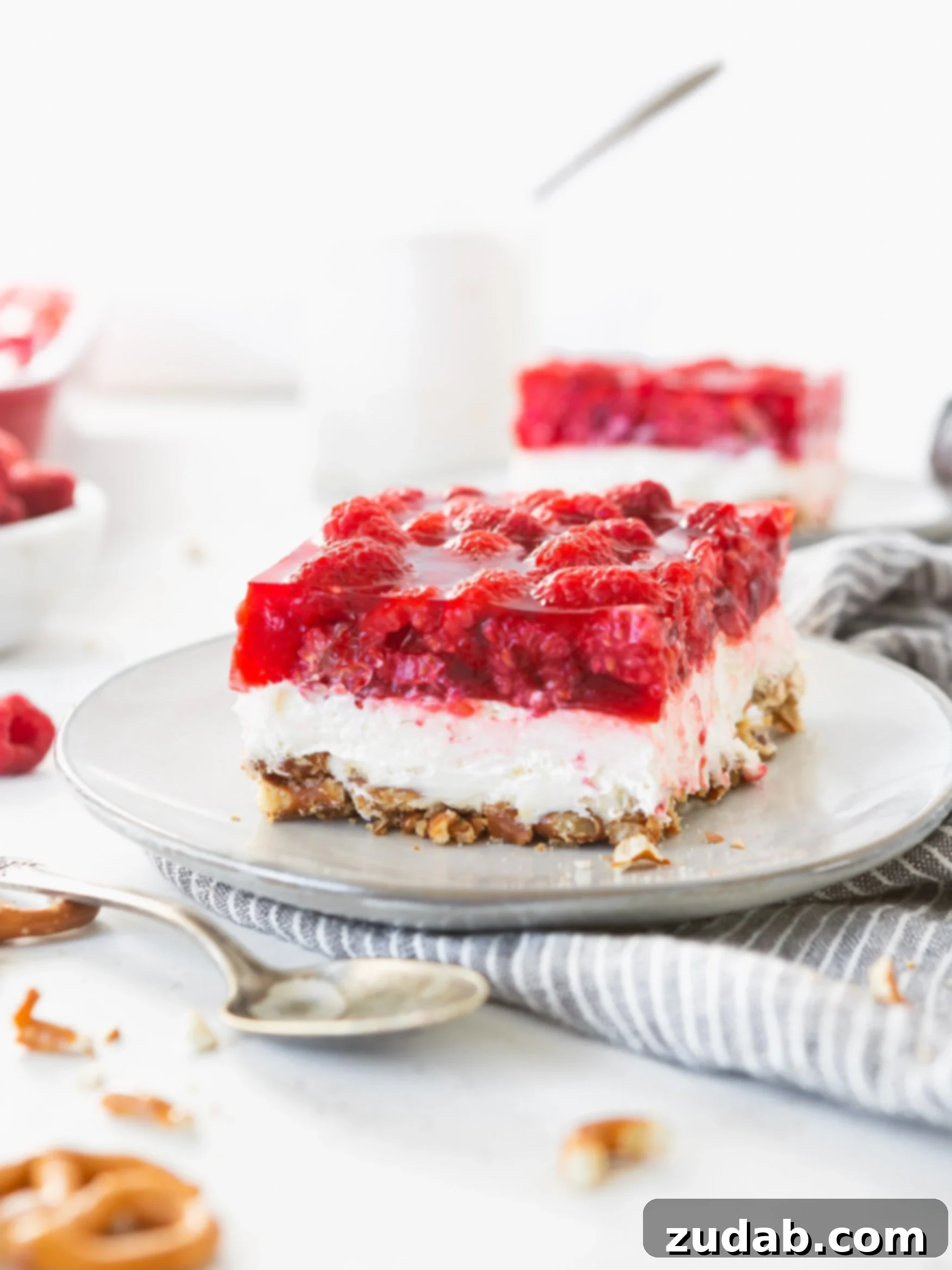 Raspberry Pretzel Jello Salad A serving of raspberry pretzel jello salad. You can see the pretzel layer on the bottom, with a white creamy layer on top of the pretzels and the red raspberry jello layer on top. There is another serving in the background and a spoon is near the plate.