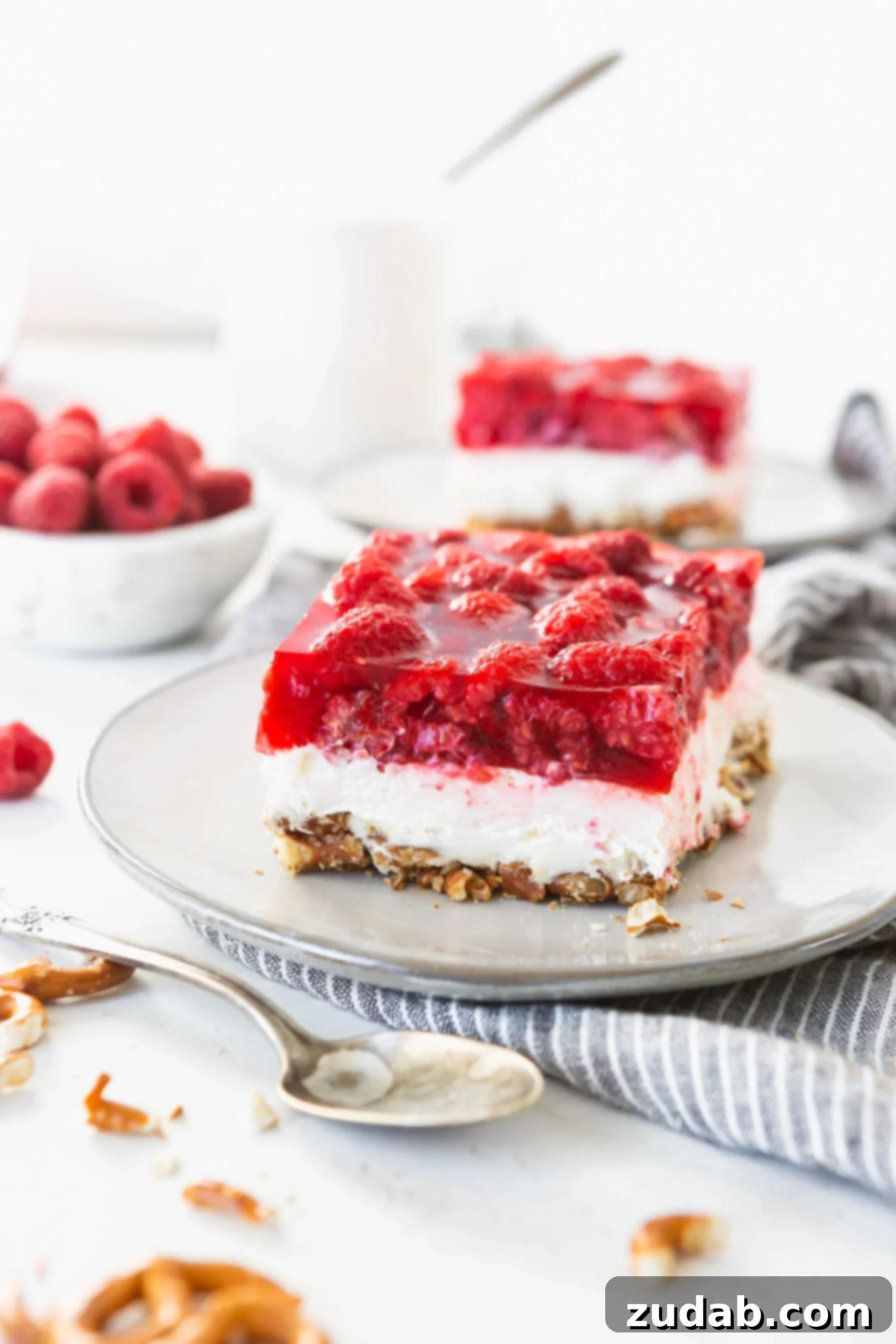 Raspberry Pretzel Jello Salad A serving of raspberry pretzel jello salad on a white salad plate. There are other servings of the salad in the background. The salad has three layers, pretzels on the bottom, a creamy layer next, and jello with fresh raspberries on top.
