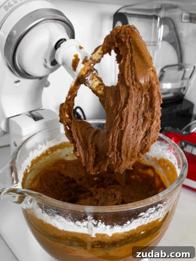 Ultimate Soft and Chewy Christmas Gingerbread Man Cookies 3 A delightful photo of soft and chewy gingerbread cookie dough filling the glass bowl of a stand mixer. The mixing paddle is lifted, showcasing the perfect consistency of the dough as it gently falls off the paddle. This dough bakes into the best Christmas cookies you will ever eat! #gingerbreadmencookies #cookierecipe #christmascookies