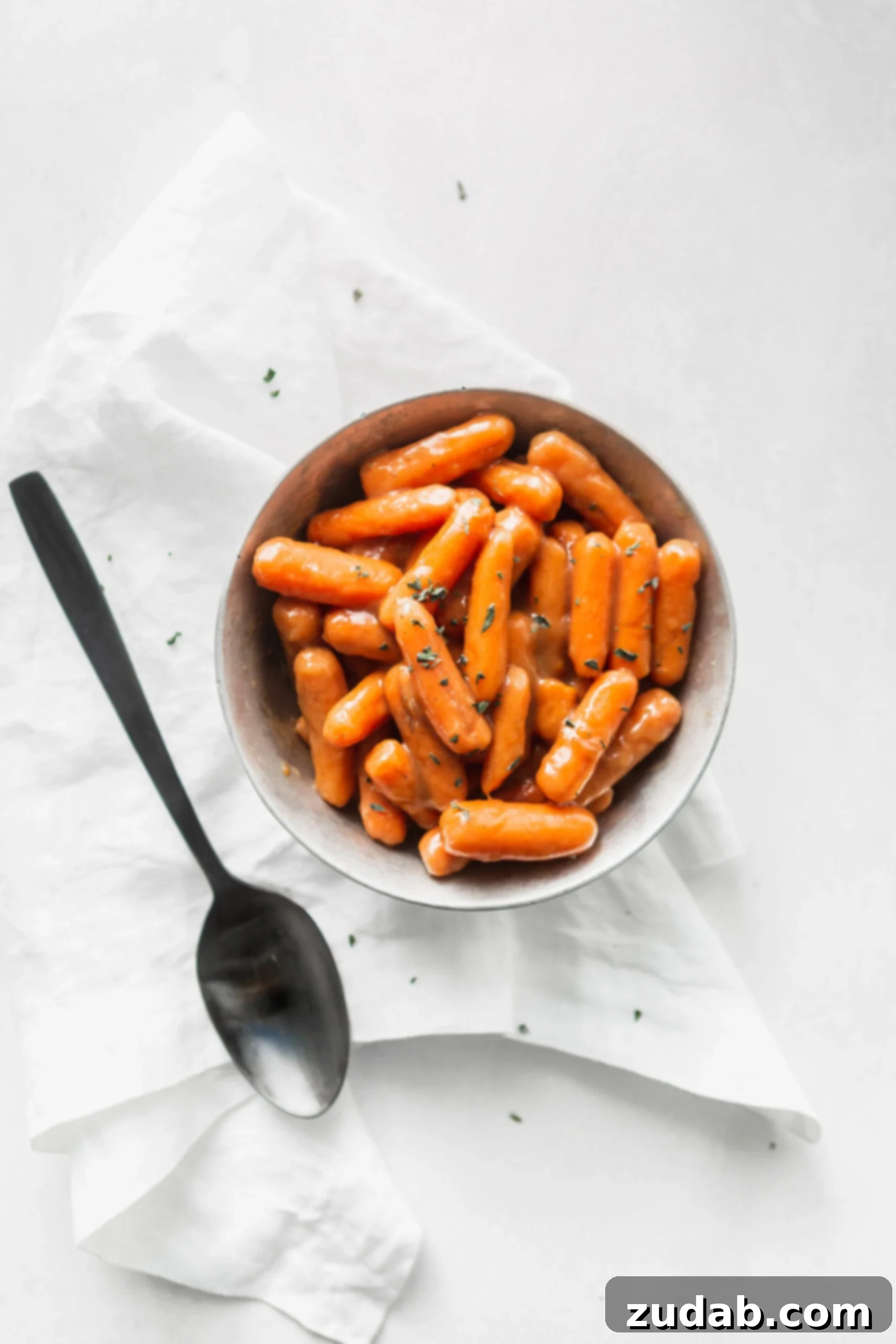 Slow Cooker Orange Glazed Carrots A serving bowl of orange glazed baby carrots. There are a few herbs sprinkled on top and a serving spoon is next to the bowl.