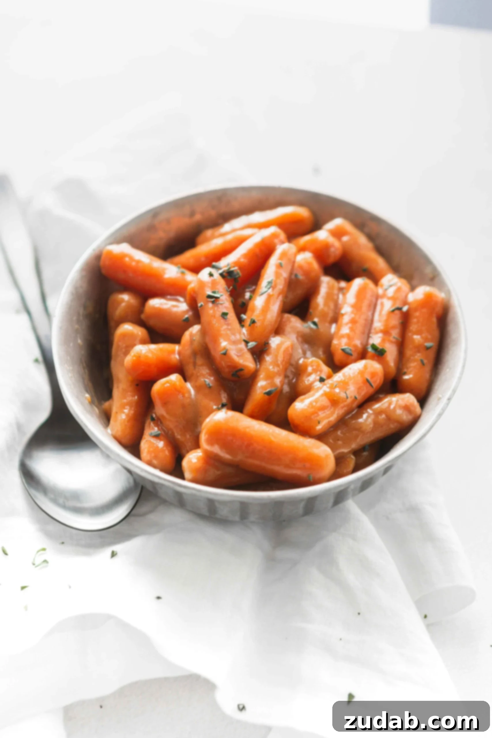 Slow Cooker Orange Glazed Carrots A bowl of orange glazed baby carrots with a little parsley sprinkled on top. A serving spoon is next to the bowl of carrots.