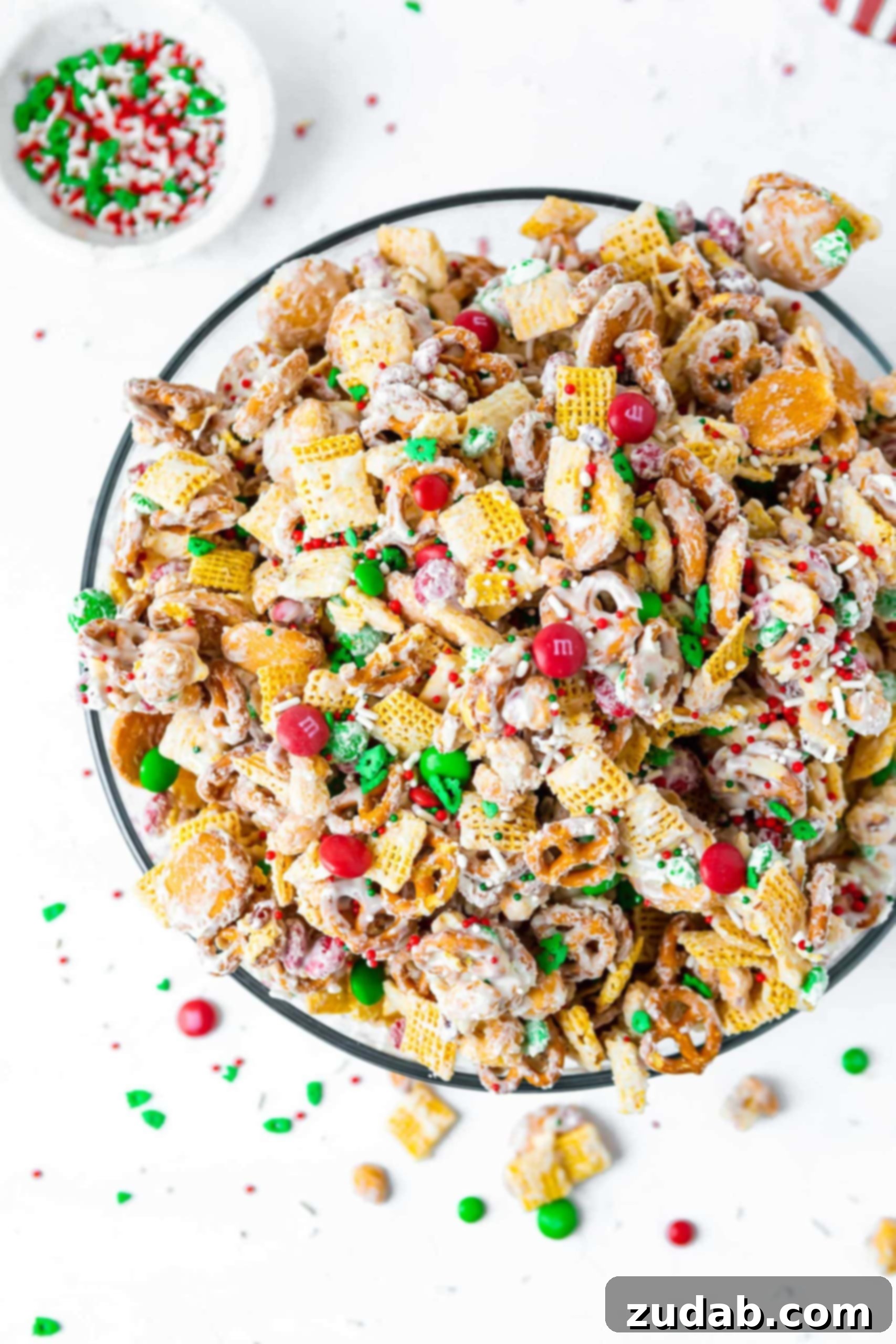 A large bowl of white chocolate Christmas Chex Mix. It contains chex cereal, pretzels, vanilla wafers, peanuts and m&m candies. It is covered in melted white chocolate chips.