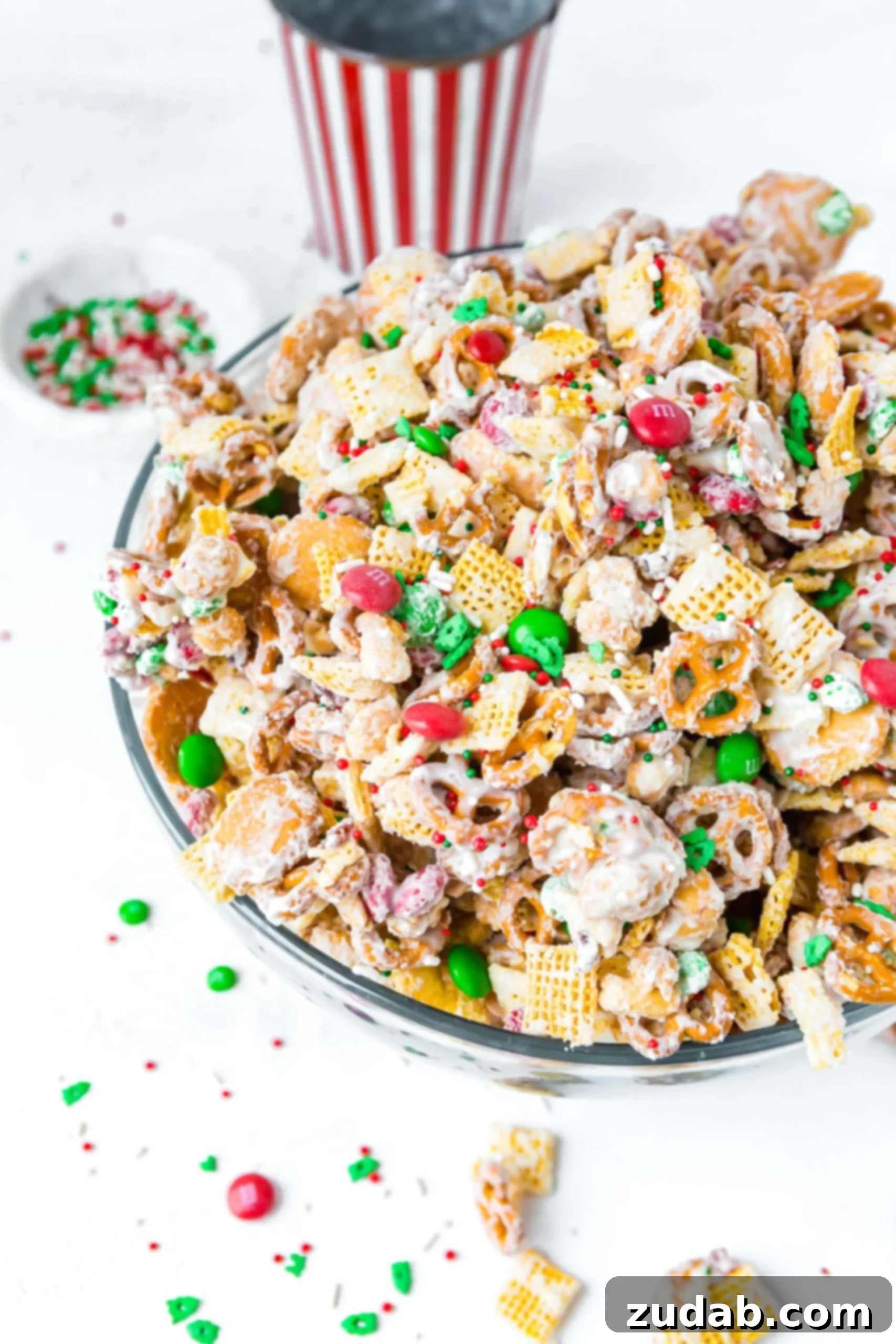 A large bowl of Christmas white chocolate trash chex mix. There are pretzels, peanuts. m&ms, chex cereal and vanilla wafers all coated with white chocolate.