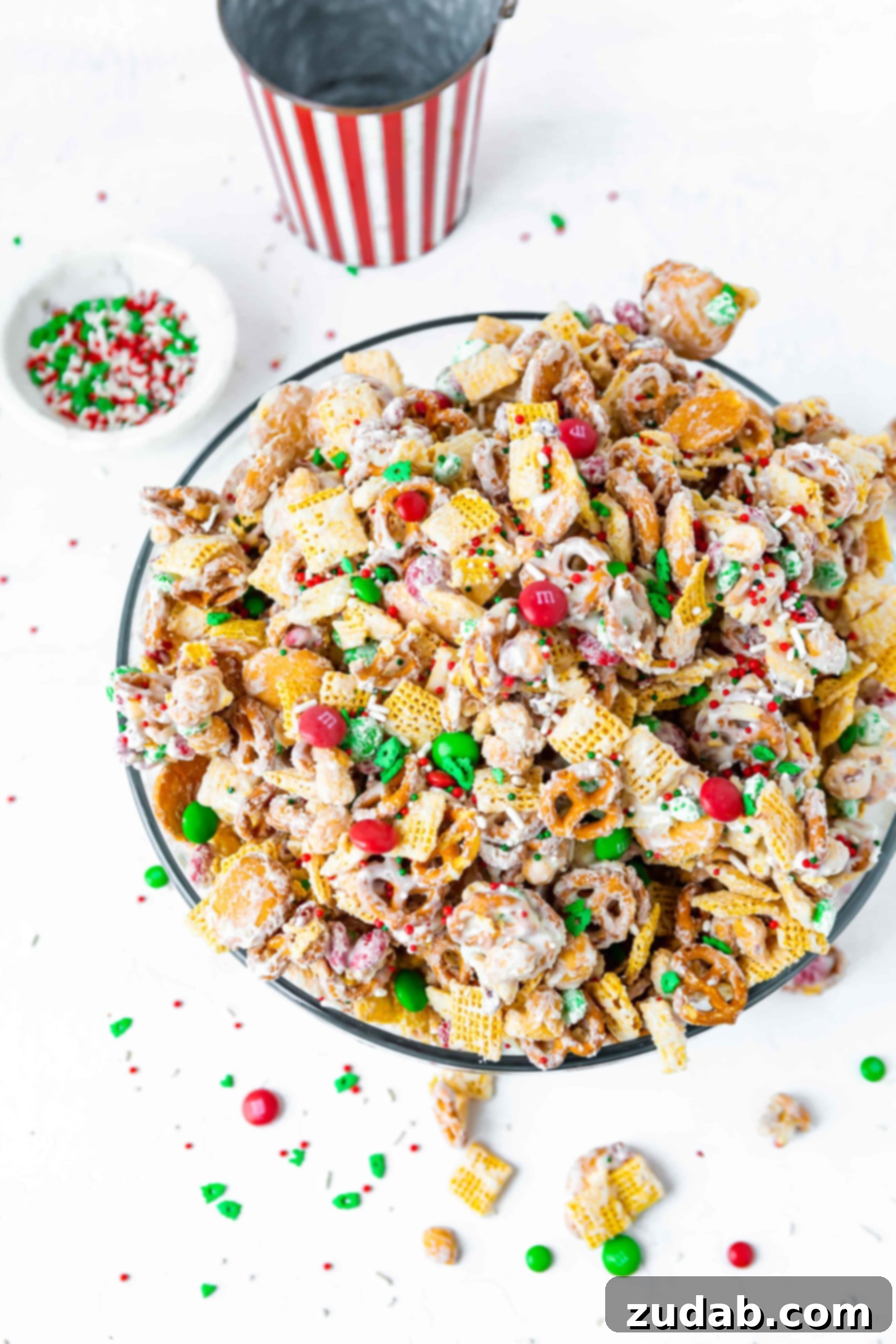 A large bowl containing Christmas white chocolate trash chex mix. It is made with two kinds of chex cereal, pretzels, vanilla wafers, peanuts and m&ms. Melted white chocolate is poured over it all.