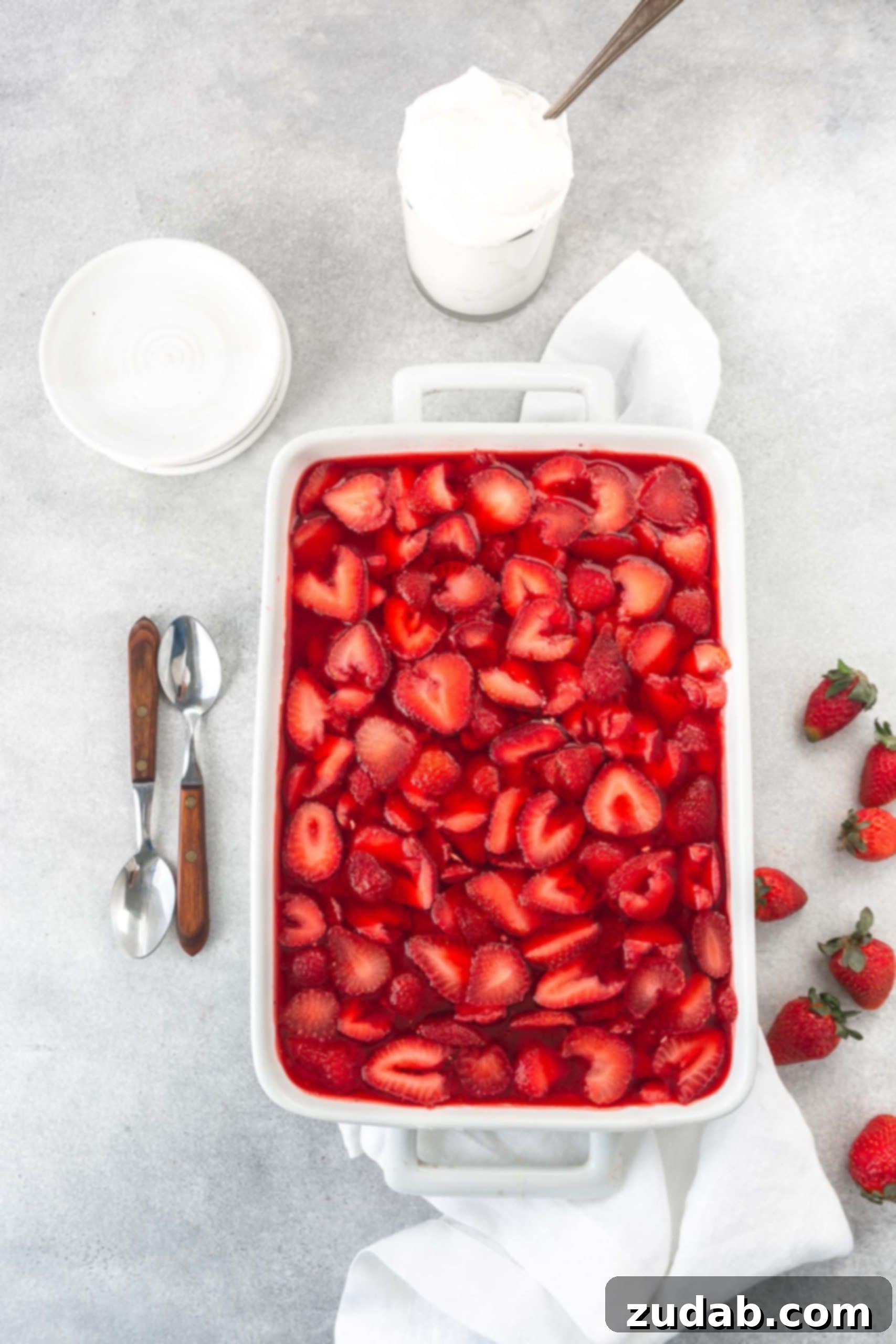 Strawberry Pretzel Dream 3 A casserole dish of strawberry pretzel jello salad. The strawberry jello is loaded with strawberry slices and there are two spoons and several strawberries beside the dish. A white bowl is in the background. The