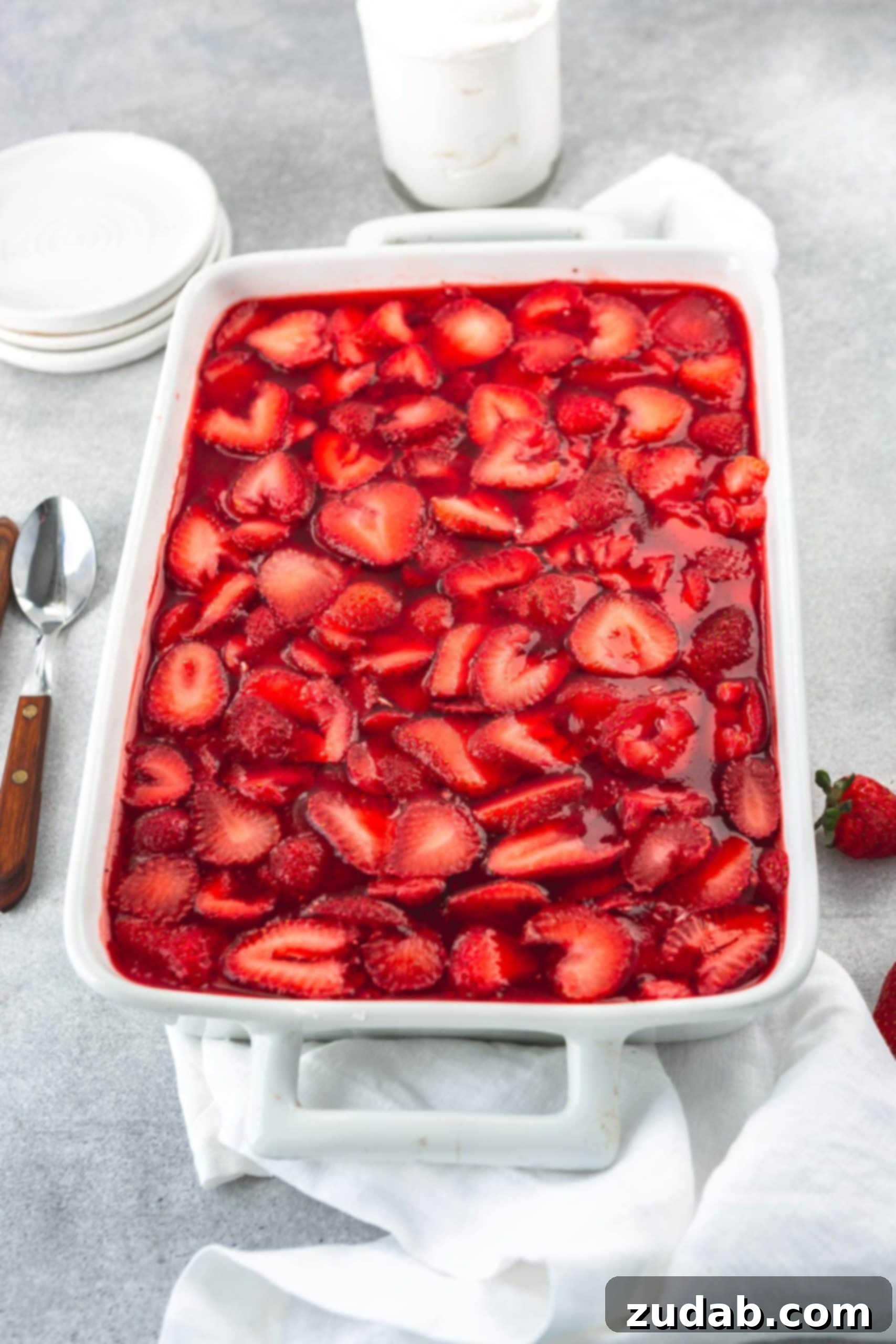 Strawberry Pretzel Dream 5 A white casserole dish with strawberry pretzel jello salad. The jello is filled with sliced strawberries and there are strawberries and two spoons next to the casserole dish.