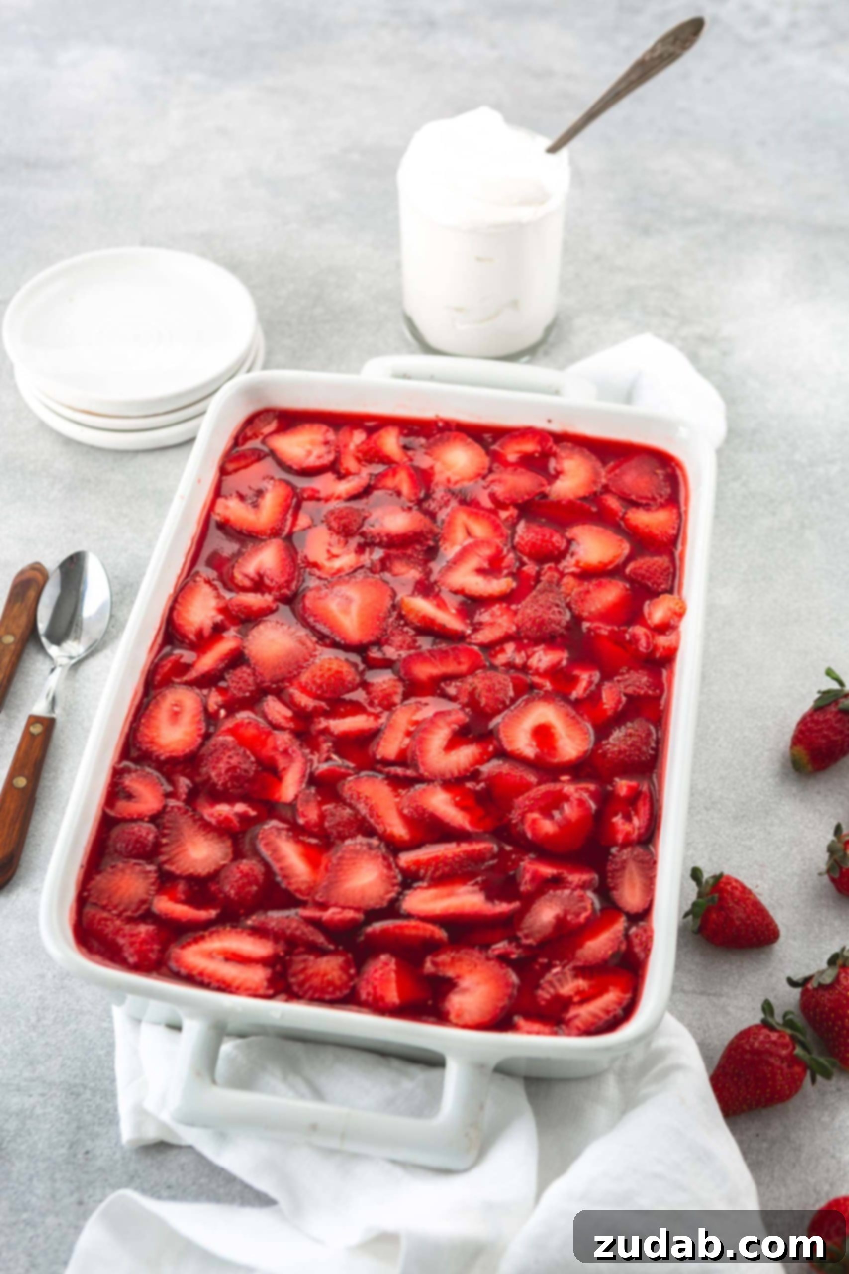 Strawberry Pretzel Dream 6 A white casserole dish with strawberry pretzel jello salad. The strawberry jello is filled with sliced strawberries. Two spoons, strawberries and a couple of small white bowls are in the background.