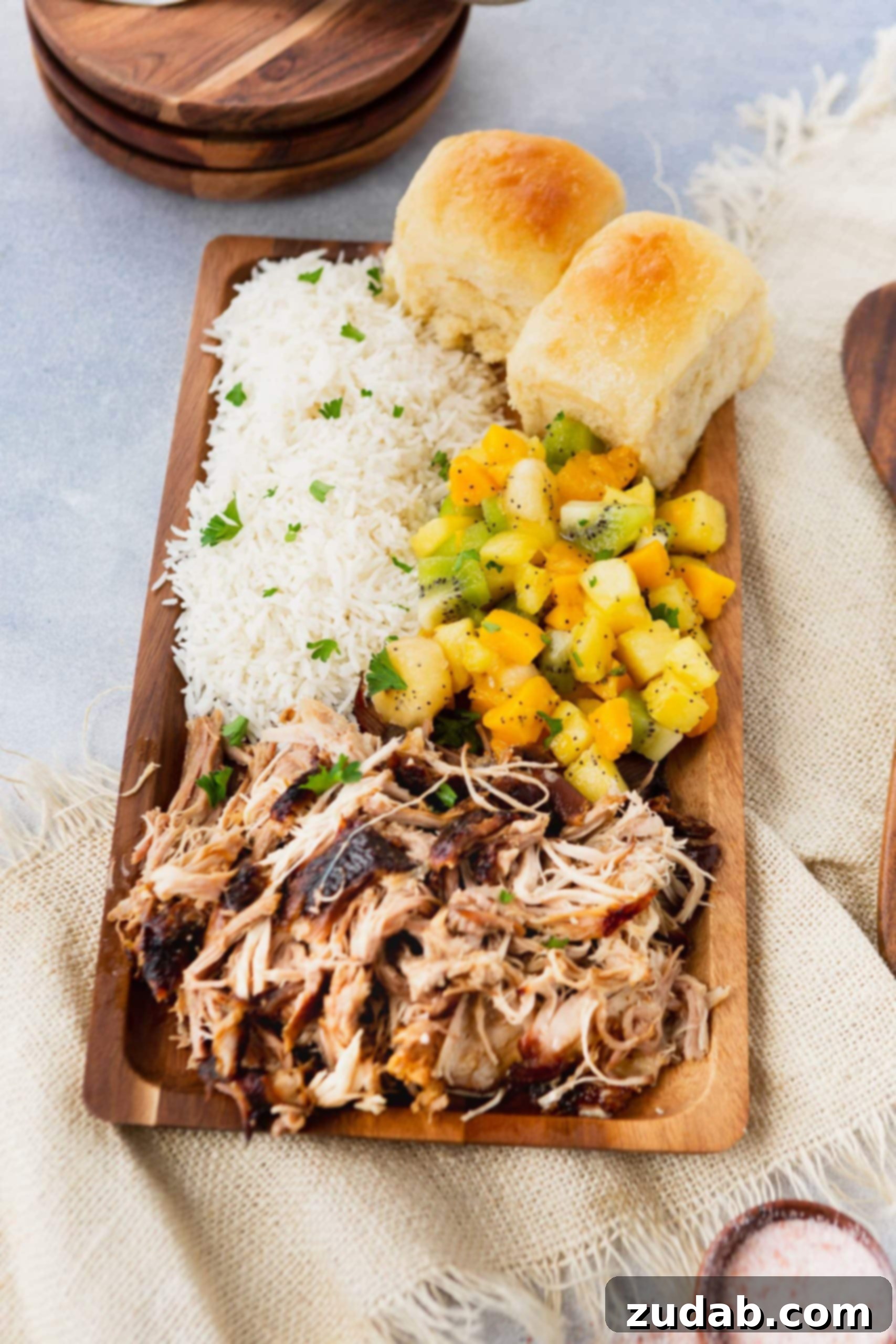 Slow Cooker Kalua Pork Recipe a photo of a rectangular wooden platter full of kalua pulled pork, herbed rice, tropical fruit salad and golden Hawaiian rolls.