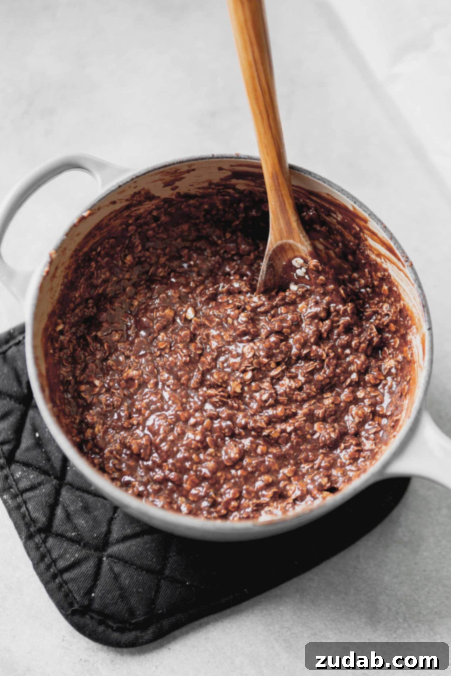 A sauce pan with no bake cookies. a wooden spoon is in the dough. You can see the oatmeal and the chocolate all mixed together.
