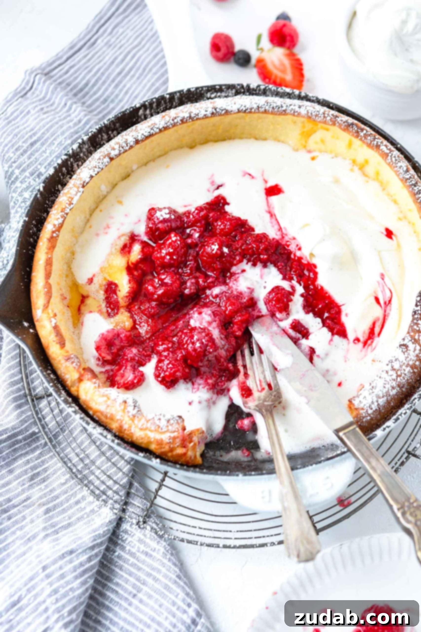 Dutch Baby Pancake A photo of a dutch baby pancake in a cast iron skillet topped with whipped cream and fresh berries with one piece removed and a fork and knife laying on the side of the pan.