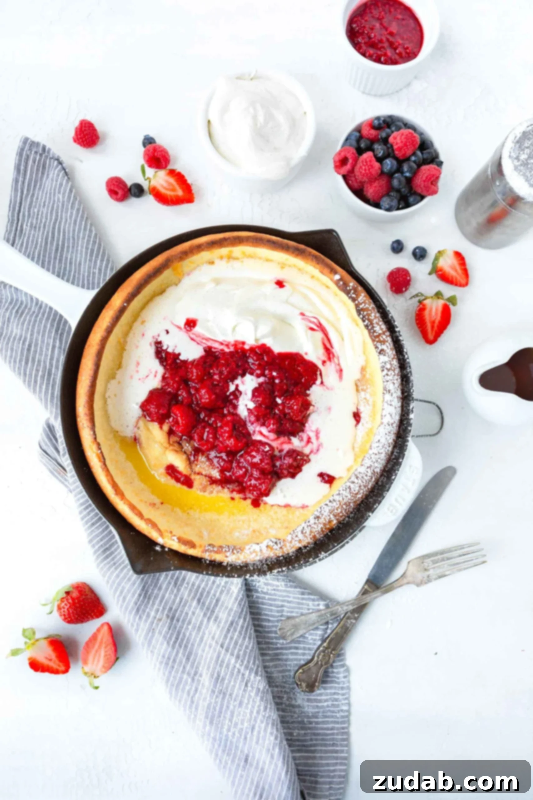 Dutch Baby Pancake A photo of a baked dutch baby pancake topped with whipped cream and fresh berries with a cup of fresh berries sitting next to the pan.