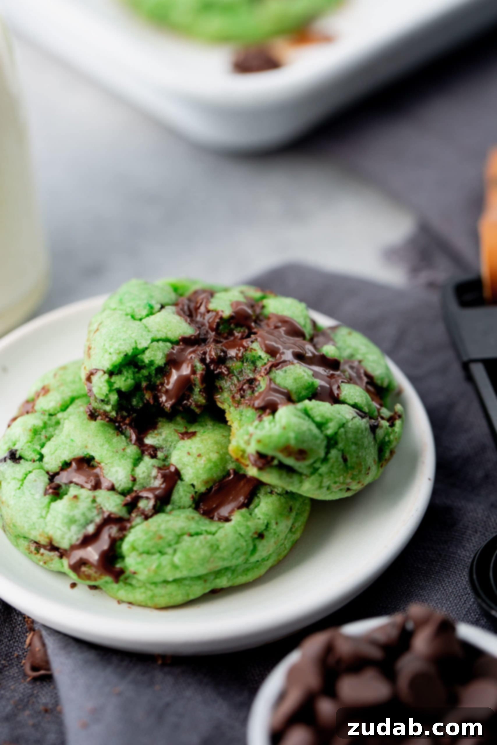 Green Mint Chocolate Chip Cookies A close-up photo of two large, soft green mint chocolate chip cookies on a small white plate. One cookie has a bite taken out of it, revealing its chewy texture and dark chocolate chips. These cookies are perfect for celebrating St. Patrick's Day with a festive and flavorful treat.