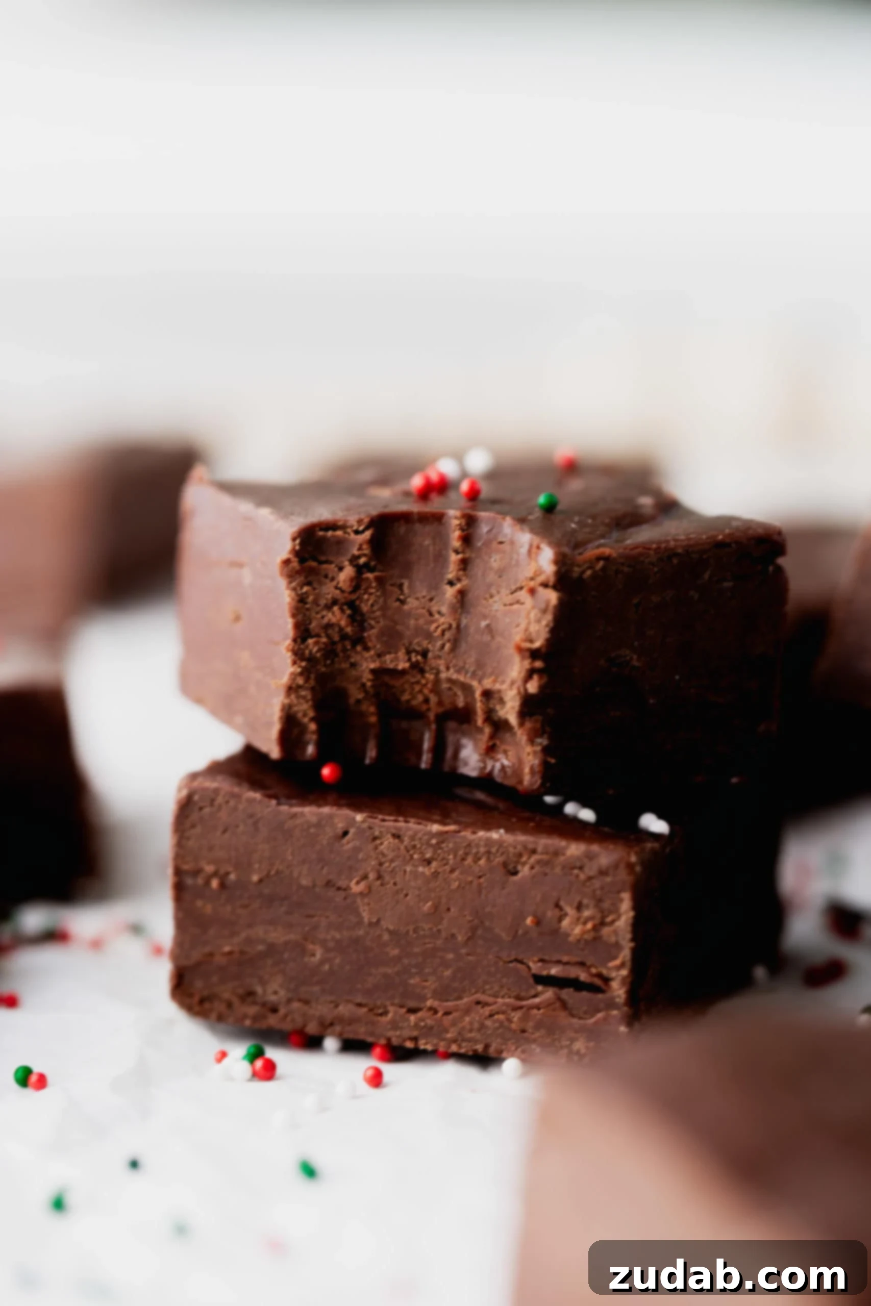 Easy Fudge Recipe Two pieces of fudge stacked on top of each other. A bite has been taken out of the top piece. A few red and green sprinkles are on and around the fudge.
