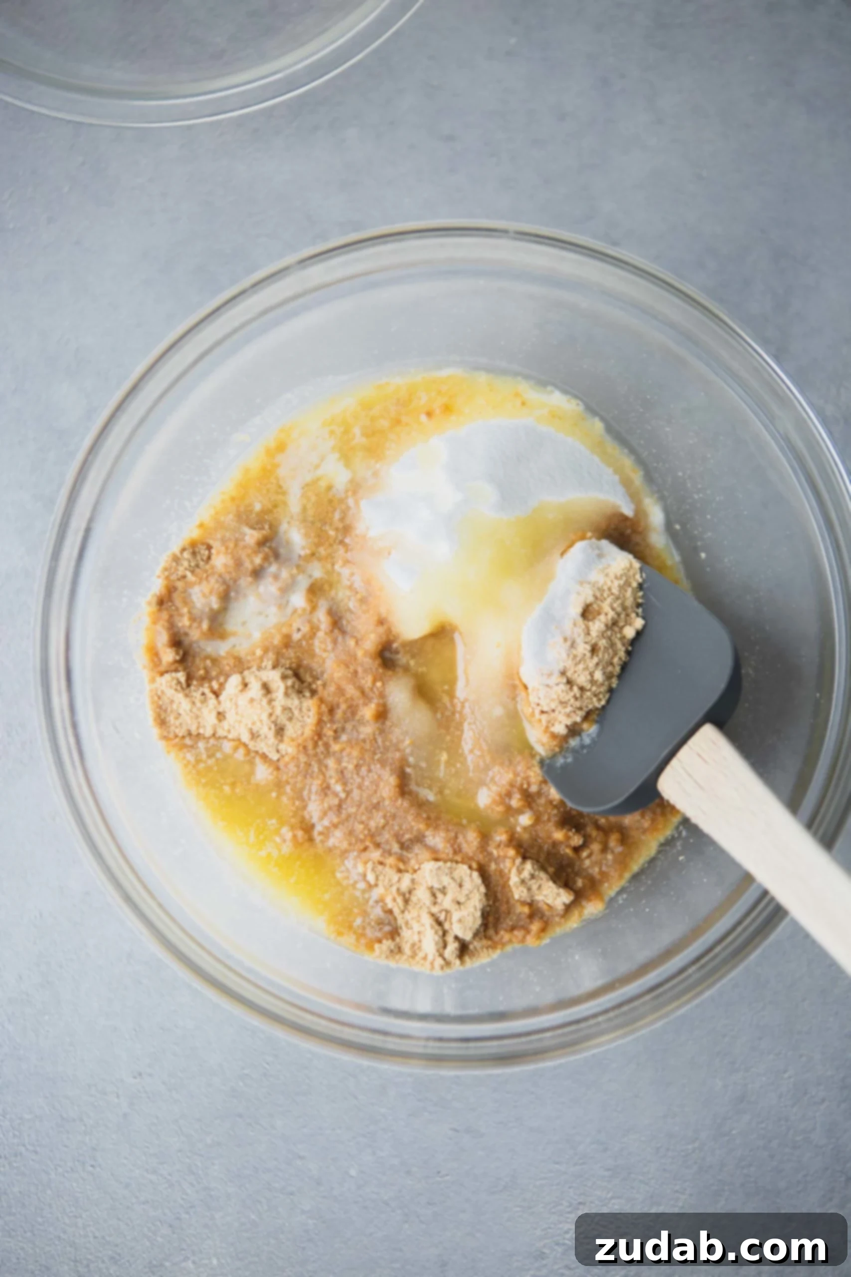 Graham Cracker Crust Ingredients Graham cracker crumbs, sugar, and melted butter are being folded together in a clear glass mixing bowl with a spatula, creating the perfect crust mixture.