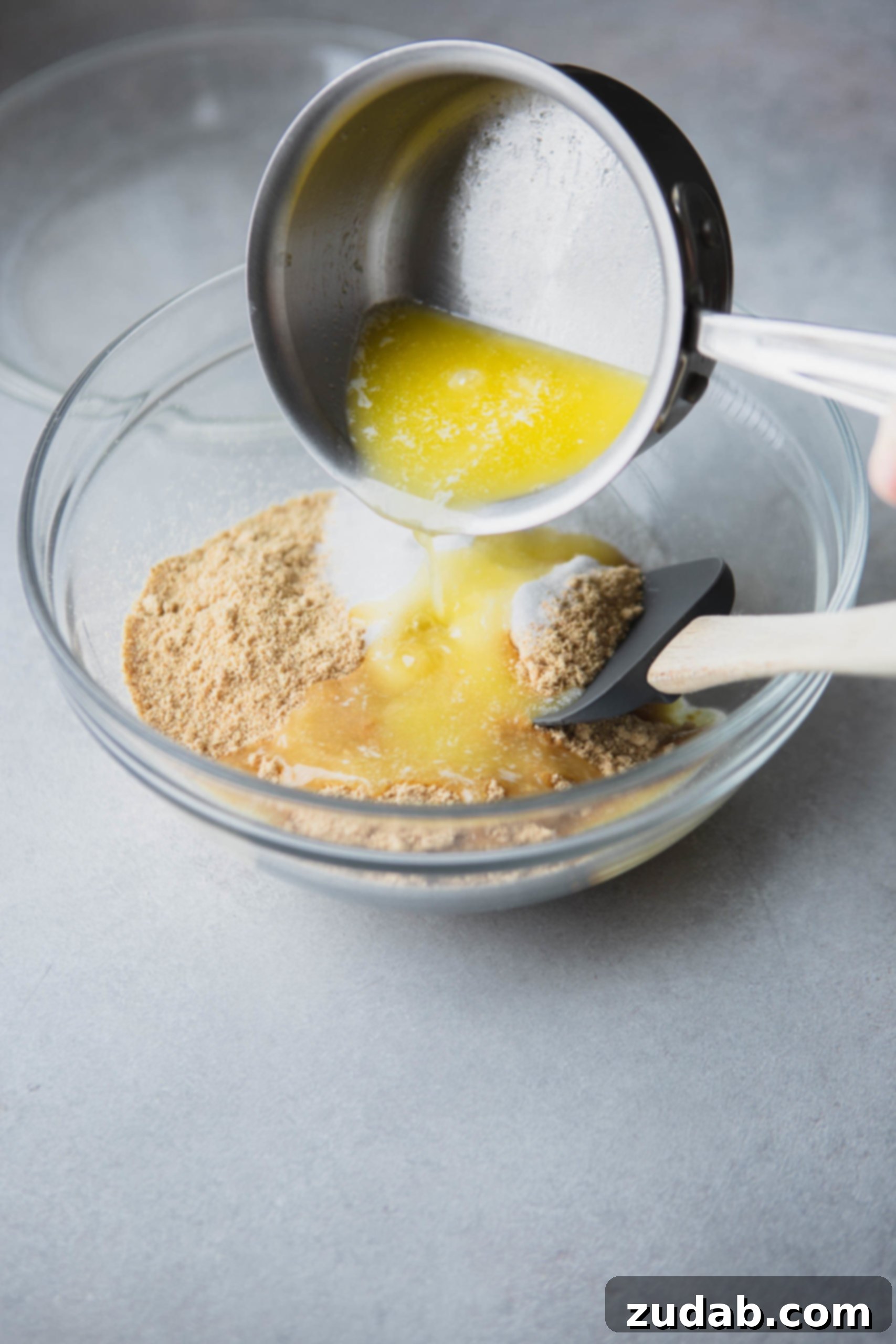 Mixing Graham Cracker Crust Ingredients Melted butter being poured over graham cracker crumbs and sugar in a clear mixing bowl, ready to be combined with a rubber spatula.