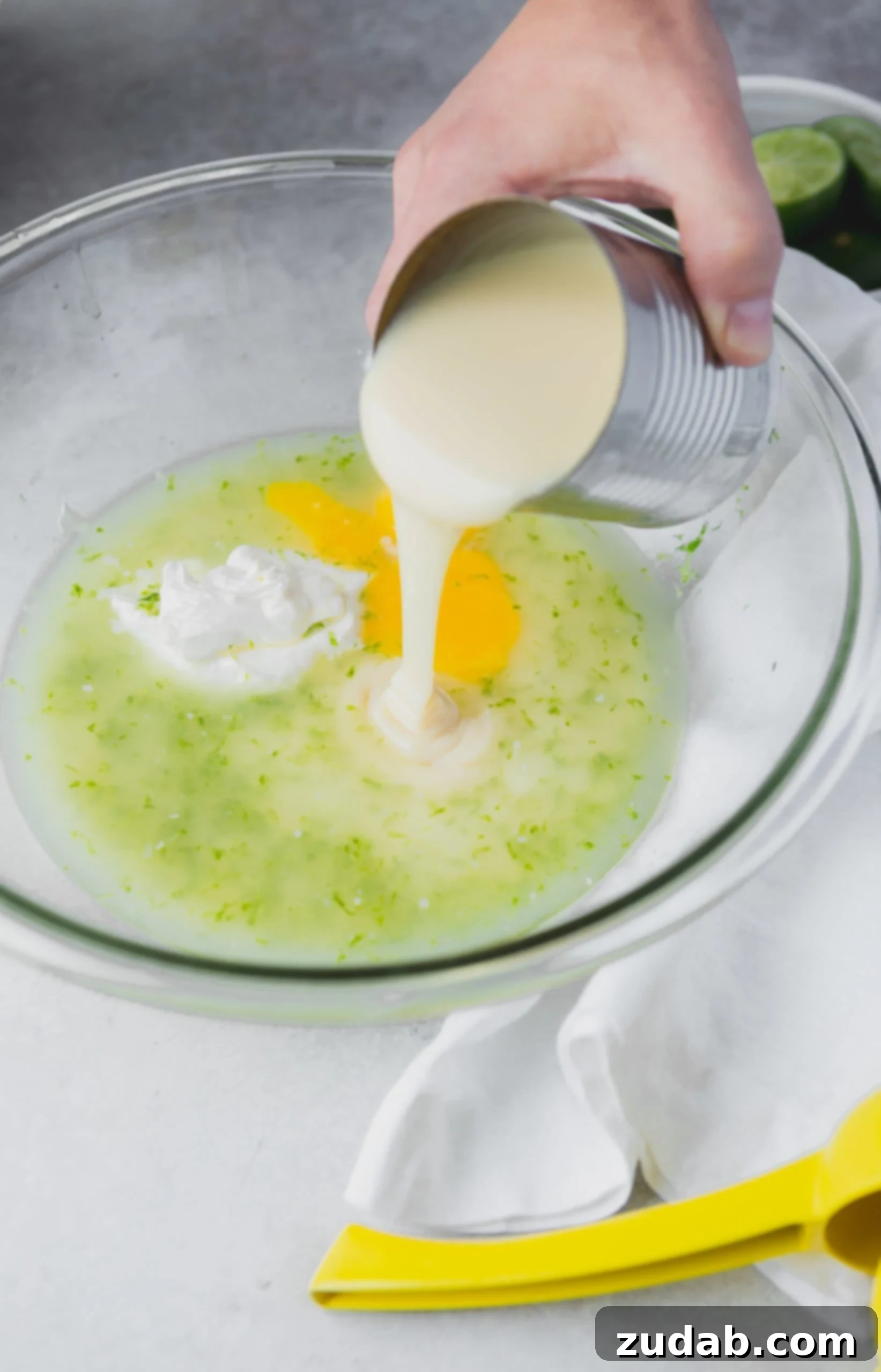 Key Lime Pie Recipe Creamy sweetened condensed milk being poured from a can into a mixing bowl with eggs, lime juice, lime zest and sour cream.