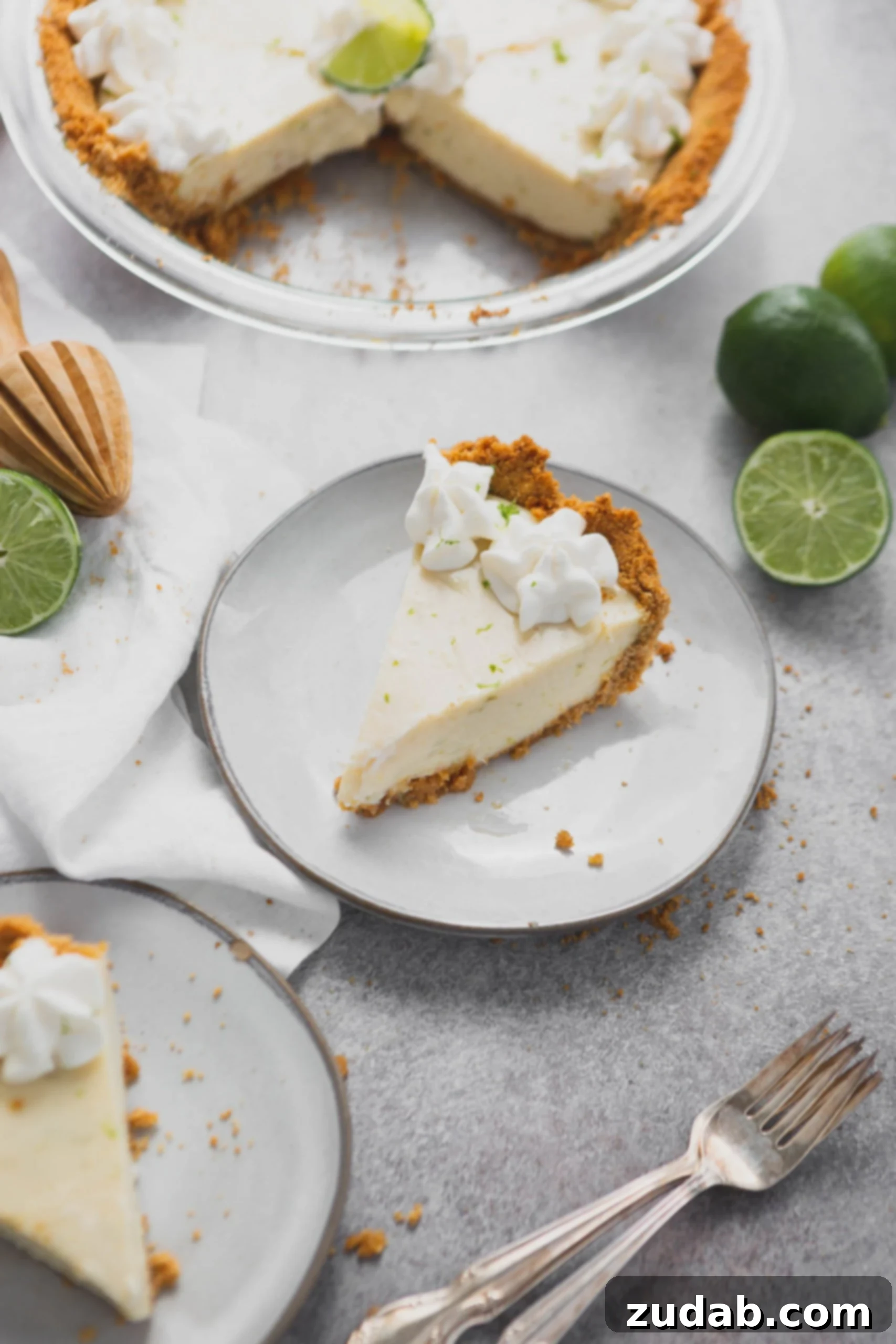 Key Lime Pie Recipe A slice of key lime pie on a white dessert plate. The graham cracker crust is filled with key lime filling and there are dollops of whipped cream on top. A lime that is cut in half is beside the plate and there is another dessert plate with a slice of pie on it.