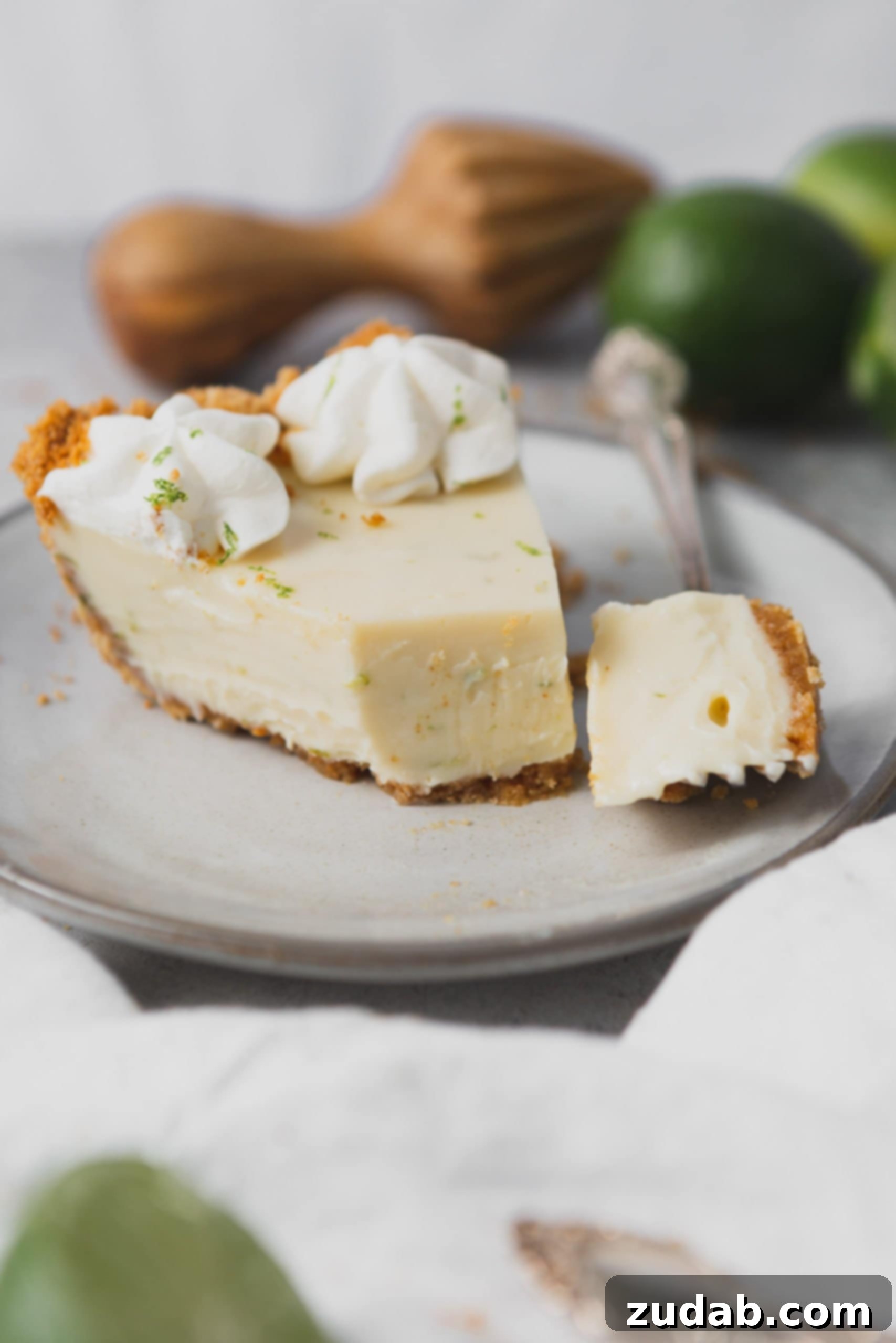 Key Lime Pie Recipe A slice of key lime pie with a piece cut from it with a fork. It is on a white dessert plate and has two dollops of whipped cream and a little lime zest on top.