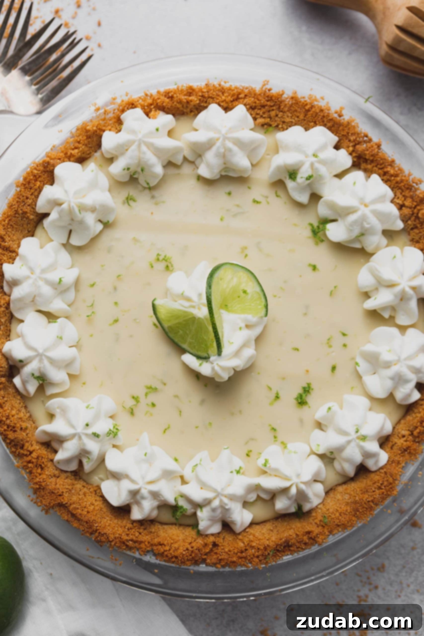 Key Lime Pie Recipe a top view of a key lime pie in a graham cracker crust. The pie has bits of lime zest, dollops of whipped cream and a curl of lime on top.