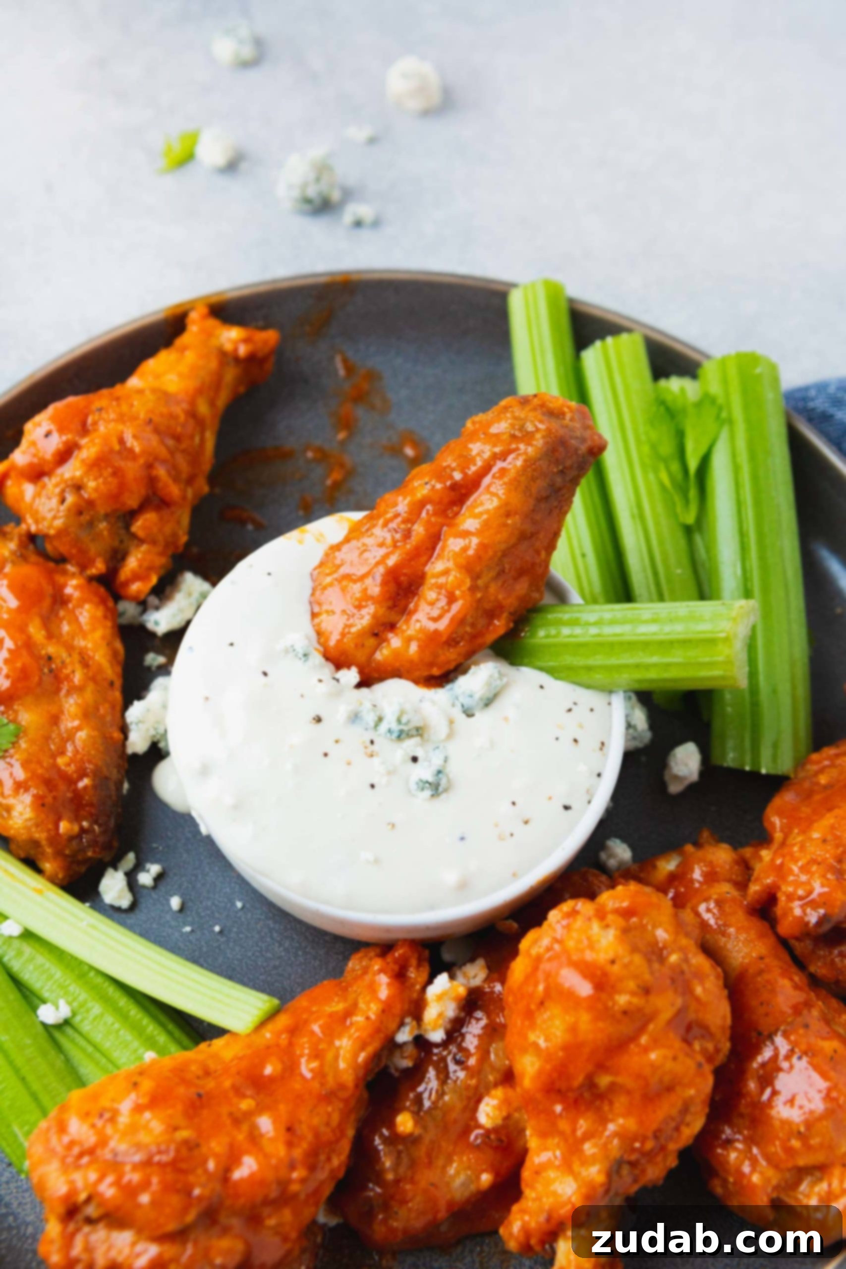 The Ultimate Crispy Baked Buffalo Chicken Wings A charcoal-colored platter featuring an abundance of perfectly cooked Buffalo chicken wings, accompanied by fresh celery sticks and a cup of creamy blue cheese dressing. One wing and one celery stick are dipped into the dressing, showcasing their readiness to be enjoyed.