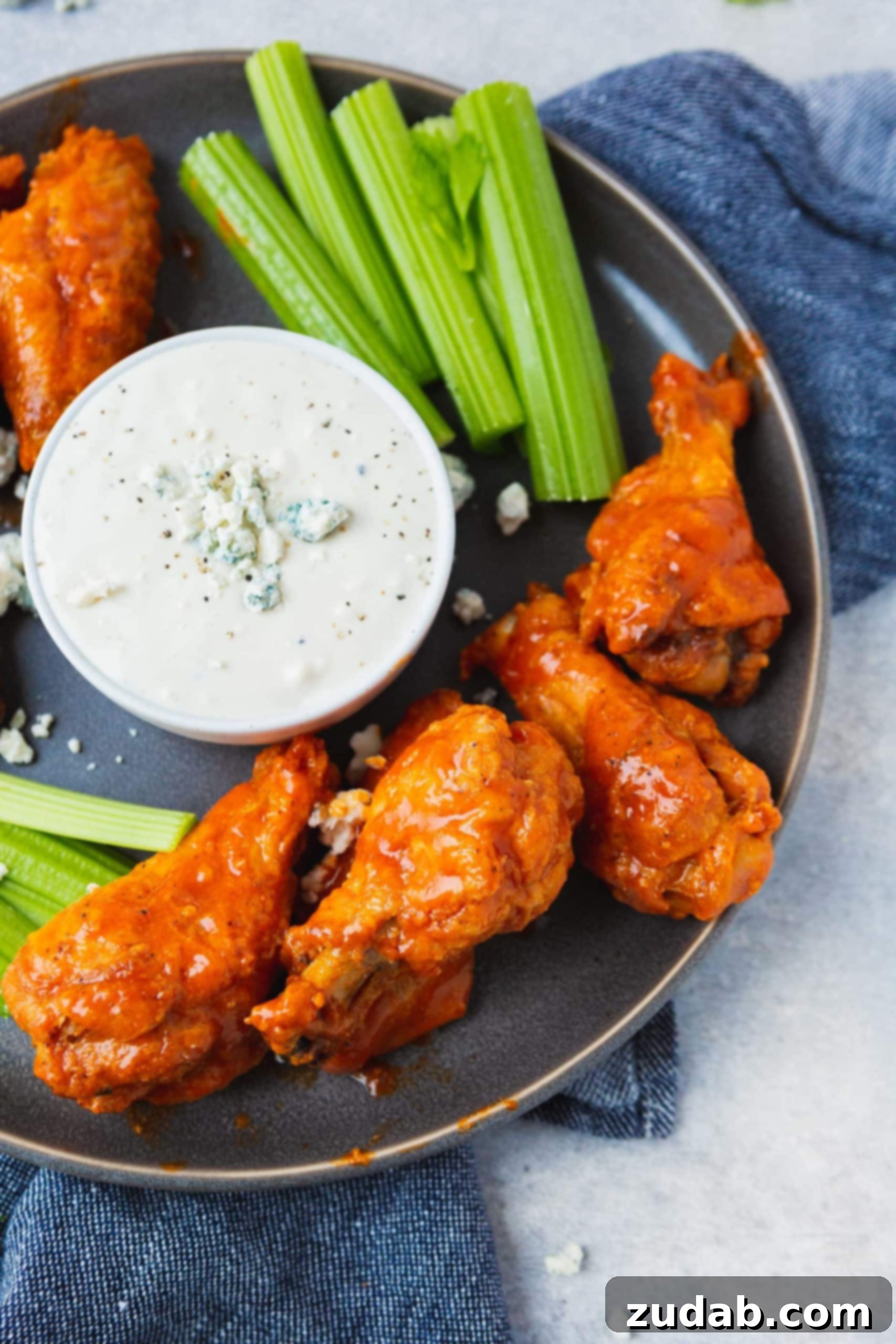 Platter of Buffalo Chicken Wings with Celery and Blue Cheese Dressing A meticulously arranged charcoal gray platter showcasing a mound of glistening, sauce-coated buffalo chicken wings surrounded by crisp celery sticks. A small cup of blue cheese dressing sits prominently in the center, inviting indulgence.