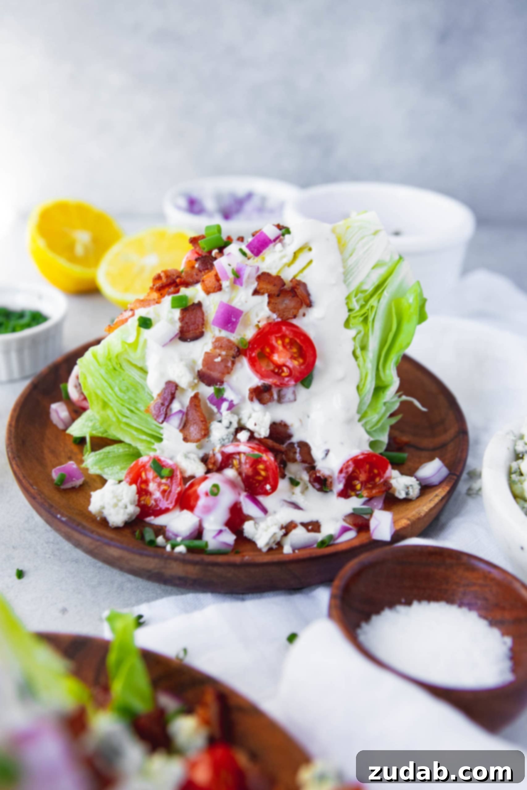 The Ultimate Wedge Salad with Bacon Lemon Drizzle a photo of a wedge salad on a wooden salad plate topped with creamy blue cheese dressing, crispy bacon crumbles, finely diced red onion, bright halved cherry tomatoes, and extra blue cheese crumbles for a rich texture and flavor contrast.