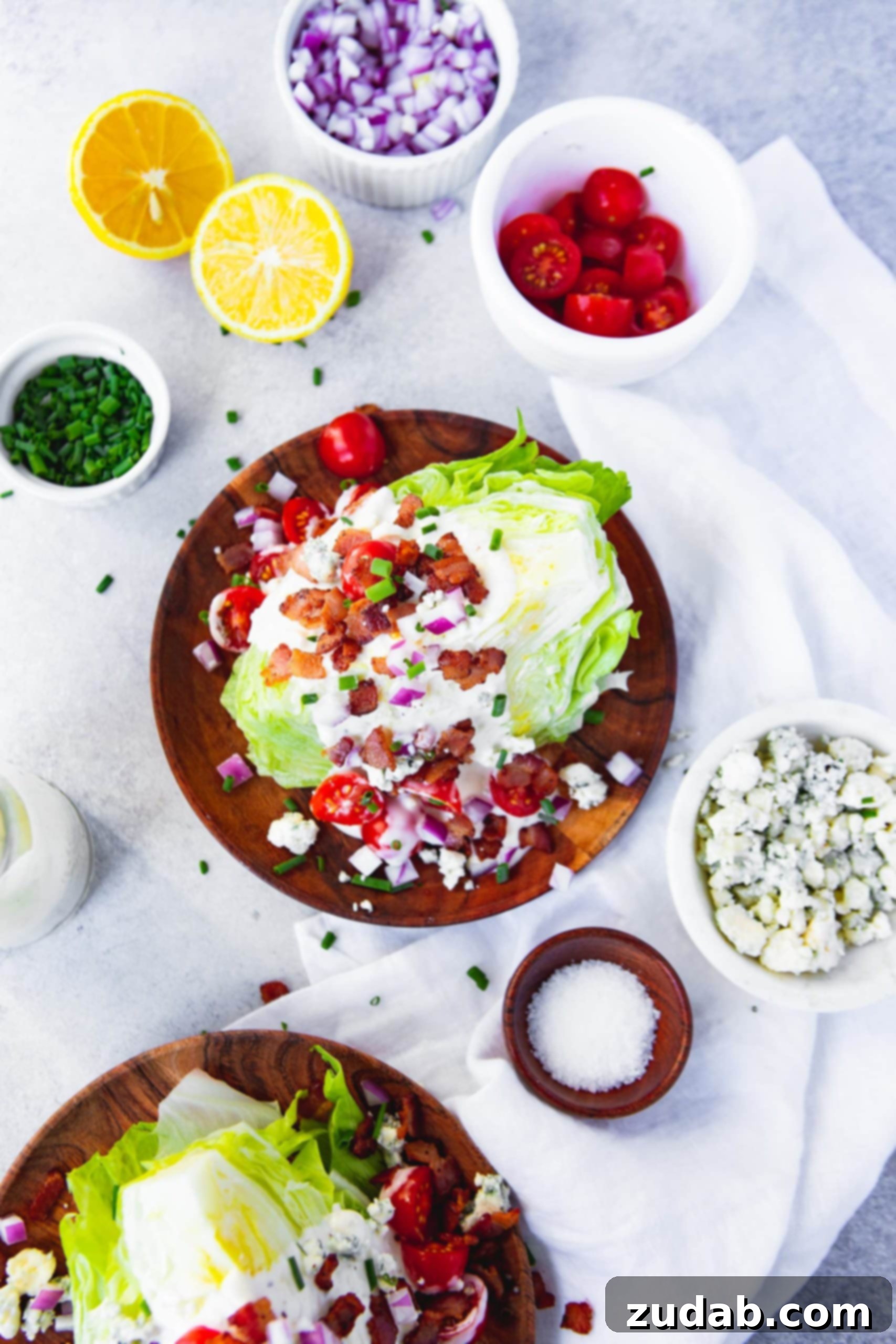 Wedge Salad with Bacon Lemon Drizzle An overhead shot of a perfectly assembled wedge salad on a rustic wooden plate. It's generously coated with creamy blue cheese dressing, crisp bacon bits, vibrant cherry tomatoes, and finely chopped red onion, surrounded by small bowls of fresh toppings.