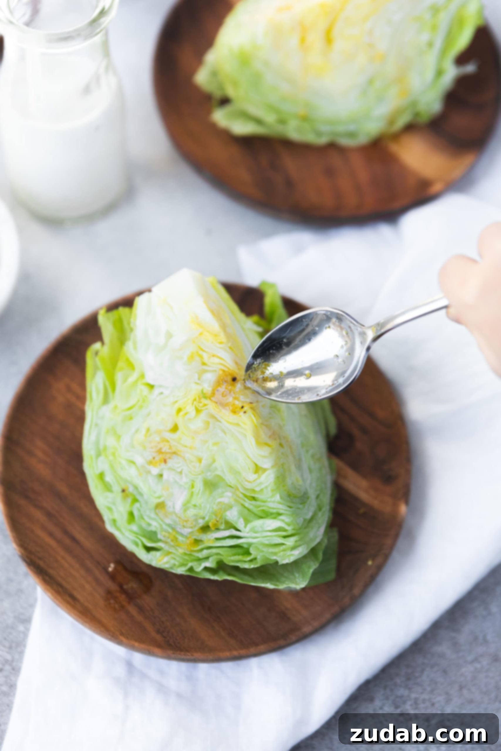 How to Make Wedge Salad a photo of a wedge of iceberg lettuce being drizzled with a bacon lemon drizzle from a silver spoon, highlighting the unique flavoring technique.