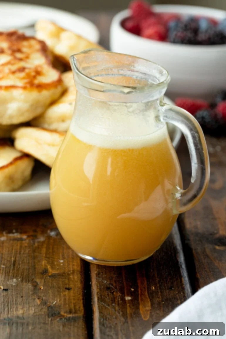 A small glass pitcher brimming with golden buttermilk syrup, set against a blurred background of fluffy pancakes, ready for a delicious breakfast.