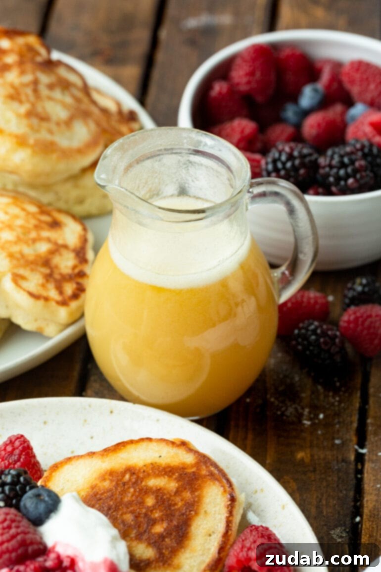 A small glass pitcher of golden buttermilk syrup is placed next to a bowl of vibrant fresh berries and a plate of golden-brown pancakes, illustrating a perfect breakfast scene.