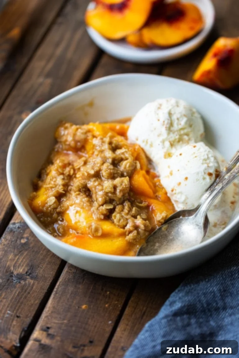Fireside Peach Crisp 2 A beautiful white bowl filled with warm, smoked fruity dessert, topped with two generous scoops of melting vanilla ice cream, ready to be enjoyed.