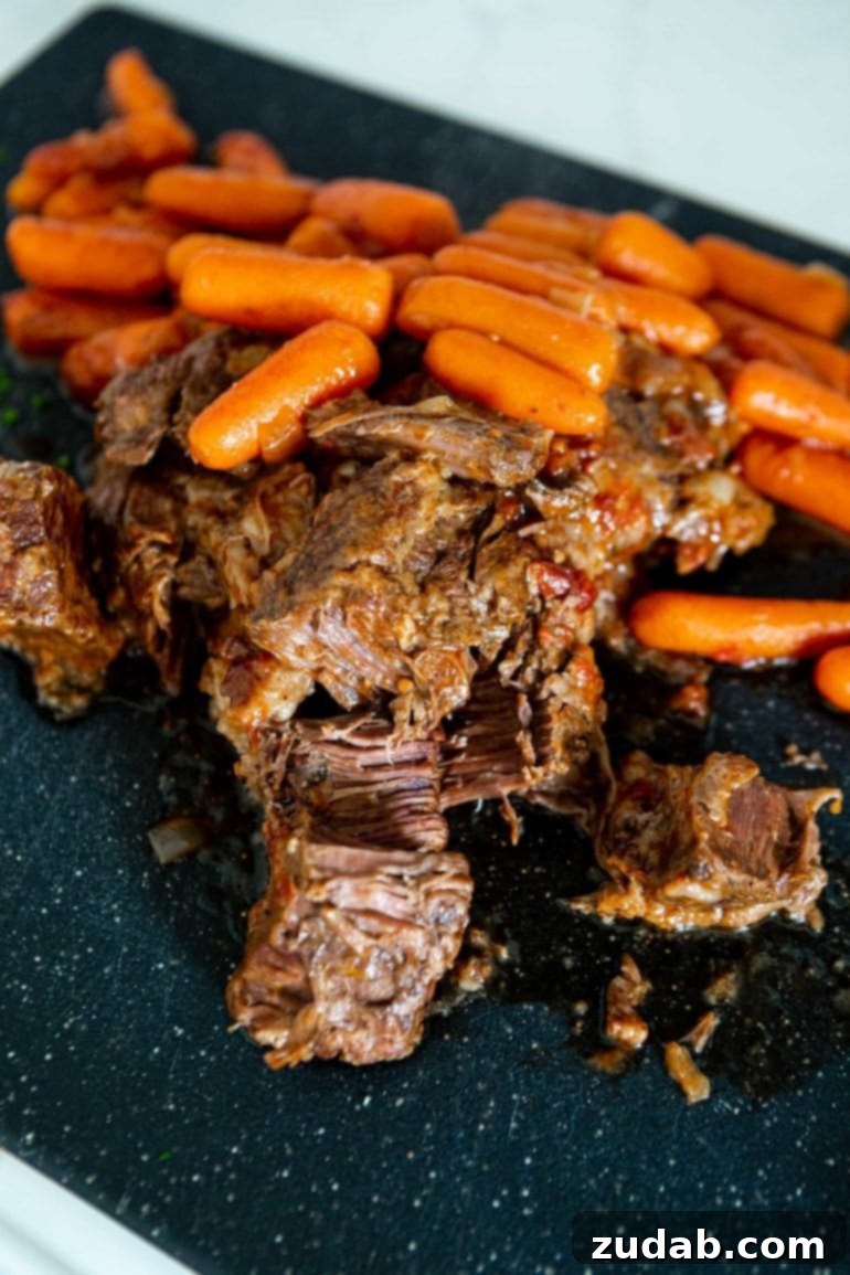 My Best-Ever Pot Roast and Gravy 3 A photo of tender beef roast being shredded on a serving platter with cooked whole baby carrots in the background.
