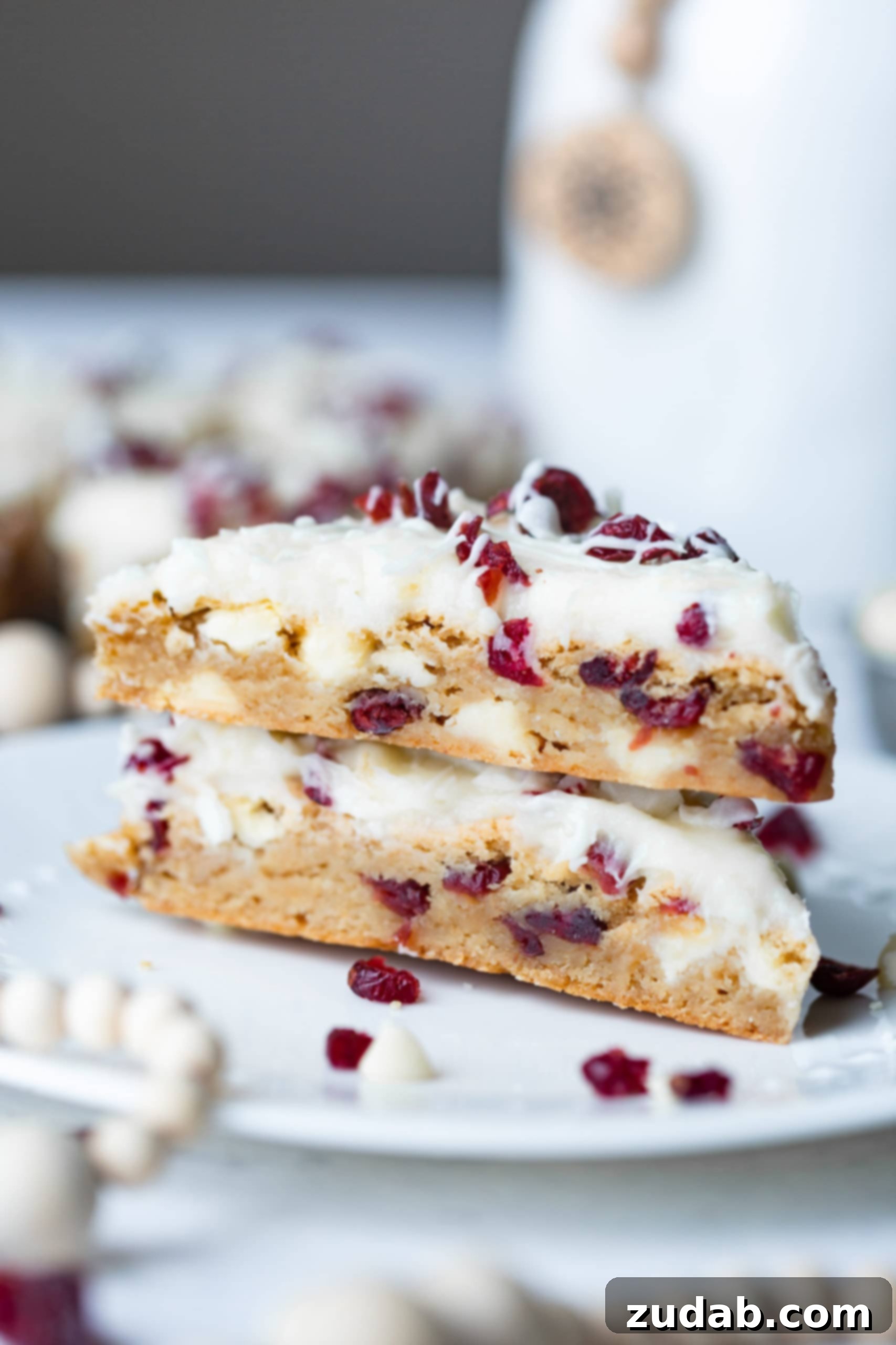 Beyaz Çikolatalı Kızılcık Keyfi 2 Two triangular white chocolate cranberry bliss bars, stacked on each other, showcasing their generous filling of white chocolate chips and craisins, topped with a smooth cream cheese frosting and a delicate white chocolate drizzle.