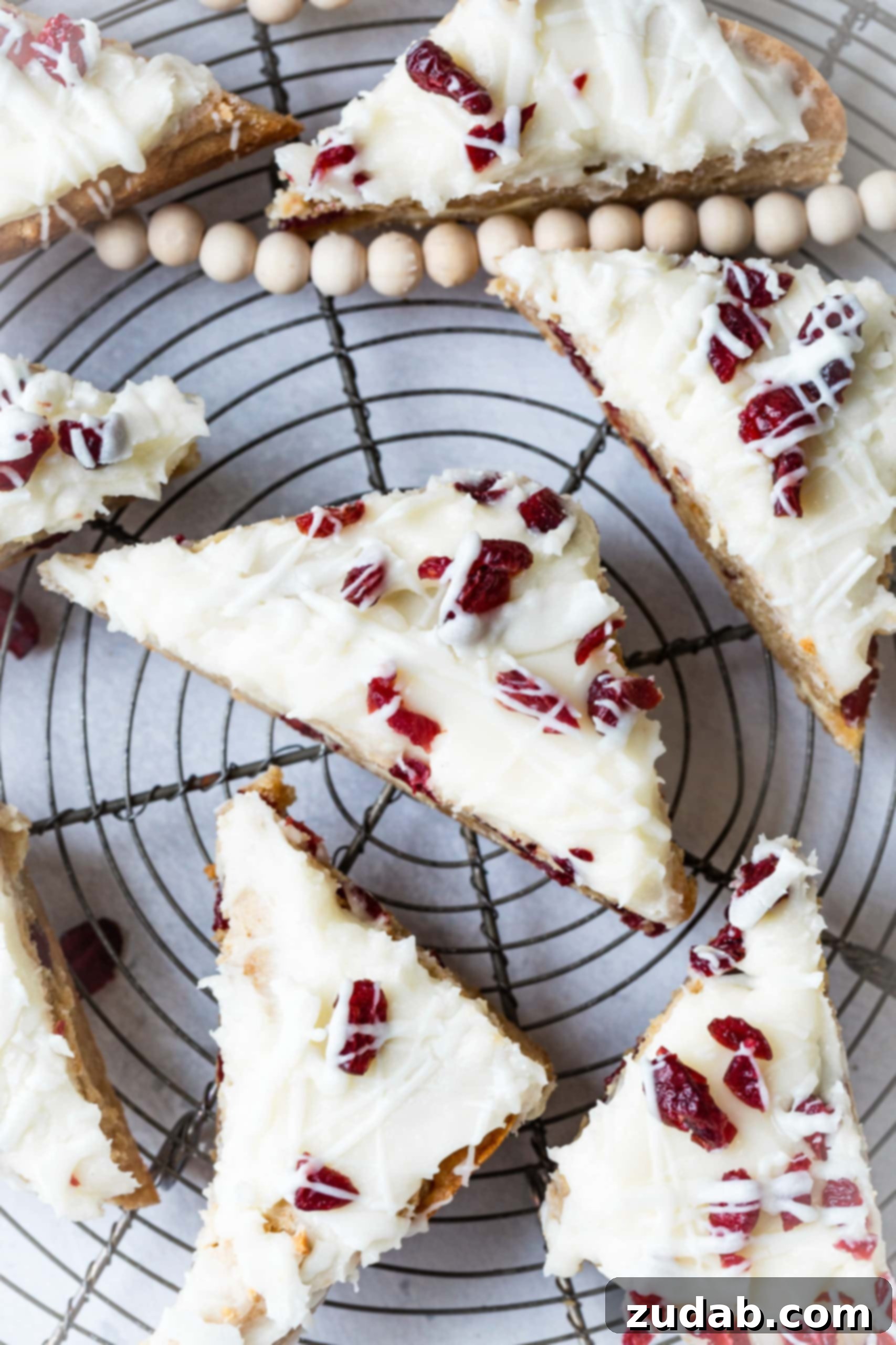 Beyaz Çikolatalı Kızılcık Keyfi 3 A photo of freshly baked white chocolate cranberry bliss bars, perfectly cut into neat triangles and arranged on a circular cooling rack, ready to be enjoyed.